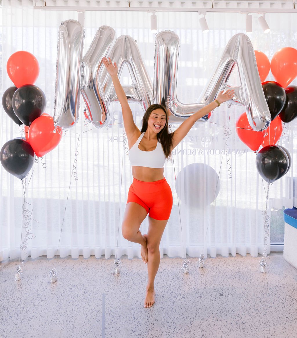A woman celebrating with arms raised in front of silver balloons spelling 'MAMA', black and red balloons, and a white curtain background.