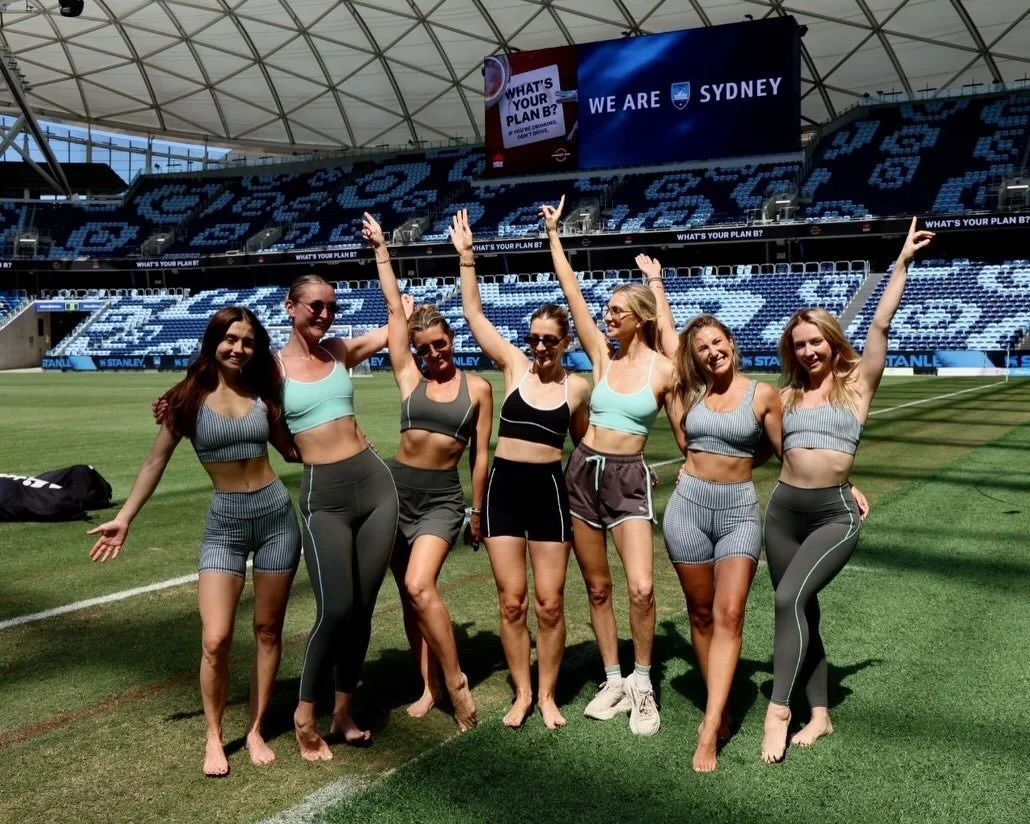 Five women in athletic wear standing with their arms raised and smiling on a sports field inside a stadium.