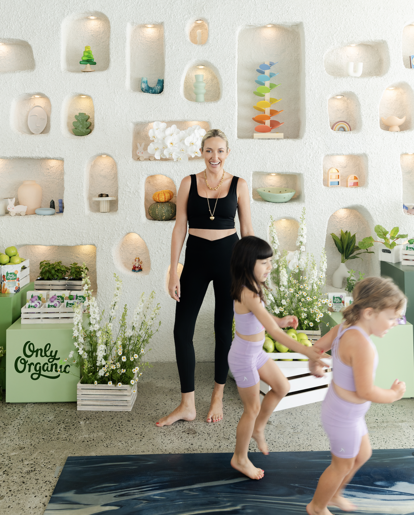 Women and children participating in a yoga class inside a store called Only Organic, with shelves of decorative ceramics, plants, and organic products on a textured white wall.