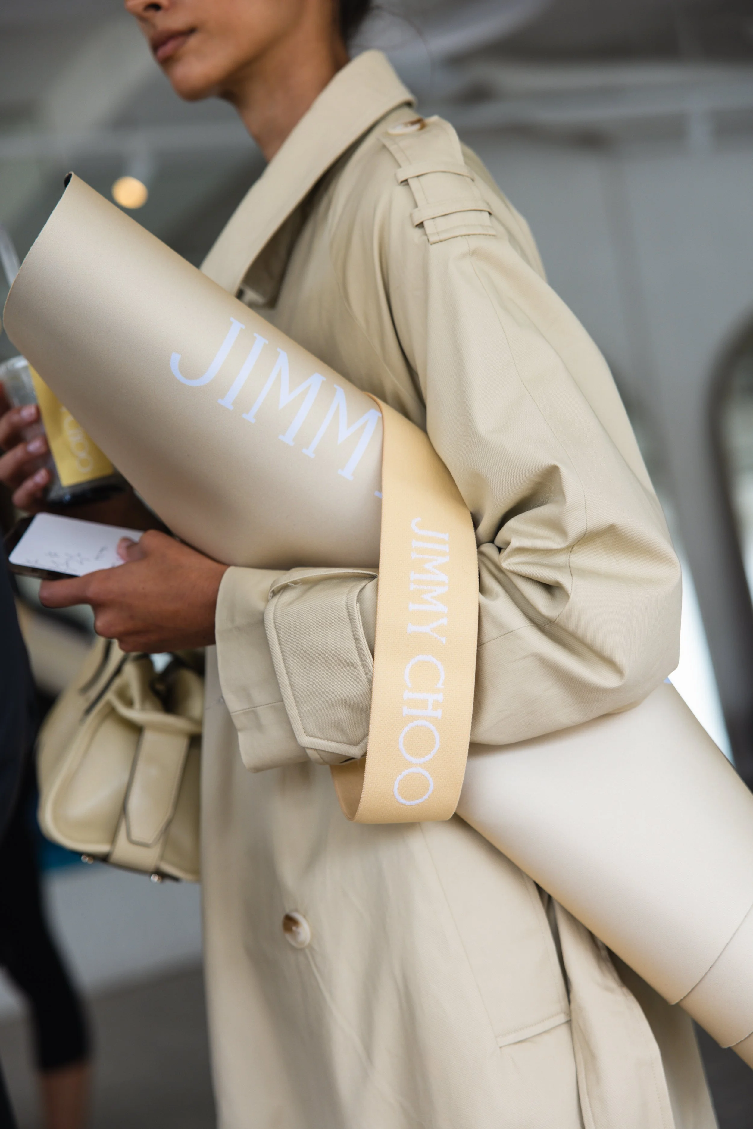 Close-up of a woman wearing a beige trench coat, holding a shopping bag with the word 'JIMMY' printed on it.