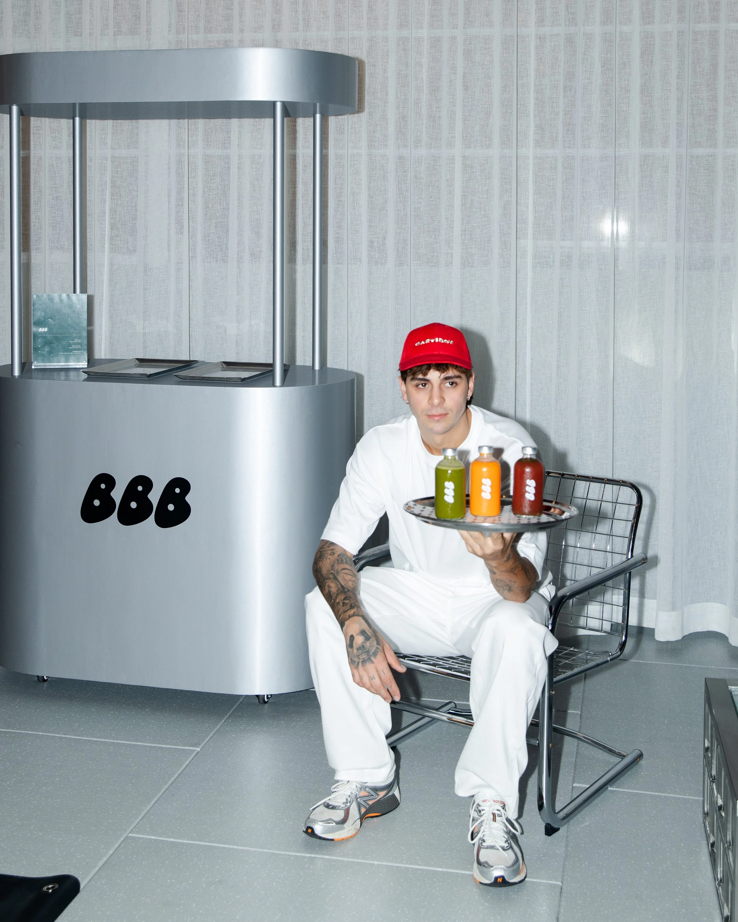 A young man wearing a red cap, white outfit, and sneakers, sitting on a metal chair, holding a tray with three bottles of different colored smoothies in front of him. Behind him is a light-colored wall and a counter with the BBC logo.
