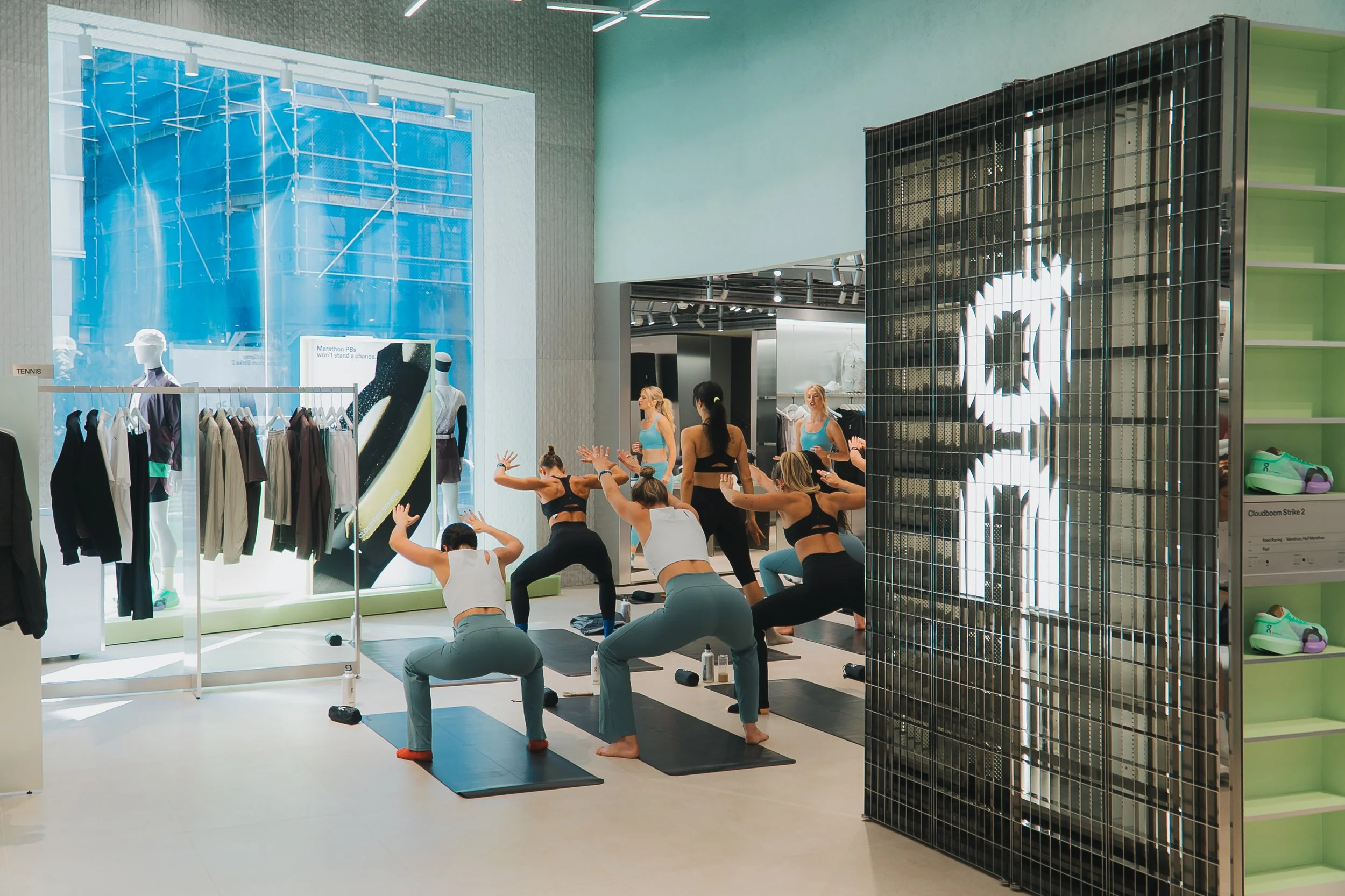 Group of women participating in a fitness class inside a retail store. They are performing squats on yoga mats, dressed in athletic wear.