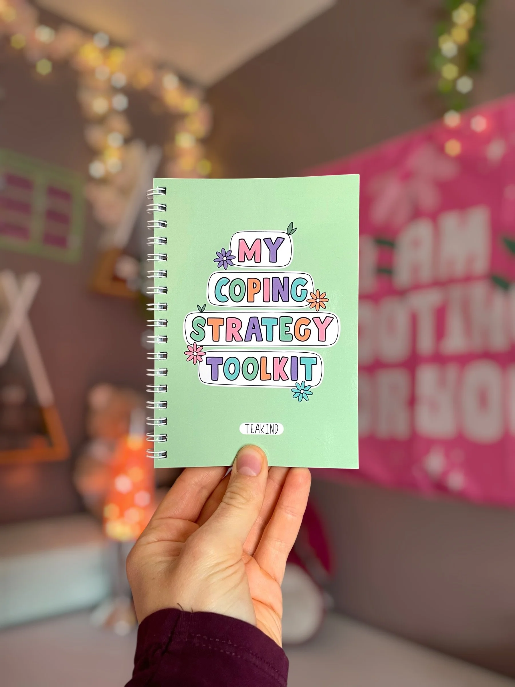 A person holding a spiral-bound notebook titled "My Coping Strategy Toolkit" with colorful decorated text and floral drawings on a green background, in a store with blurred decorations and pink packaging in the background.