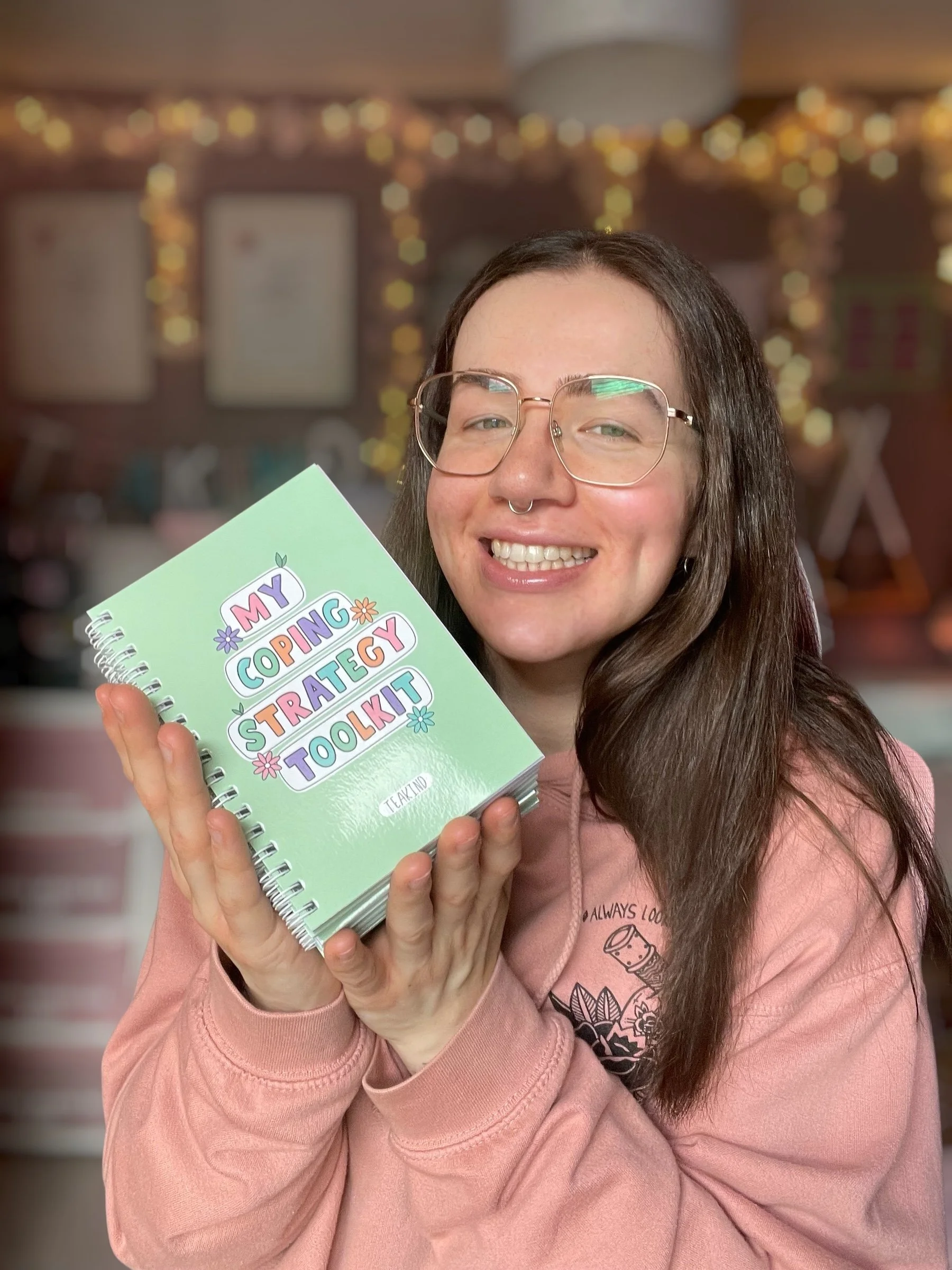 A smiling woman with glasses and a nose septum ring holds a small spiral-bound booklet titled 'My Coping Strategy Toolkit' while standing in a room decorated with blurred lights and pictures in the background.