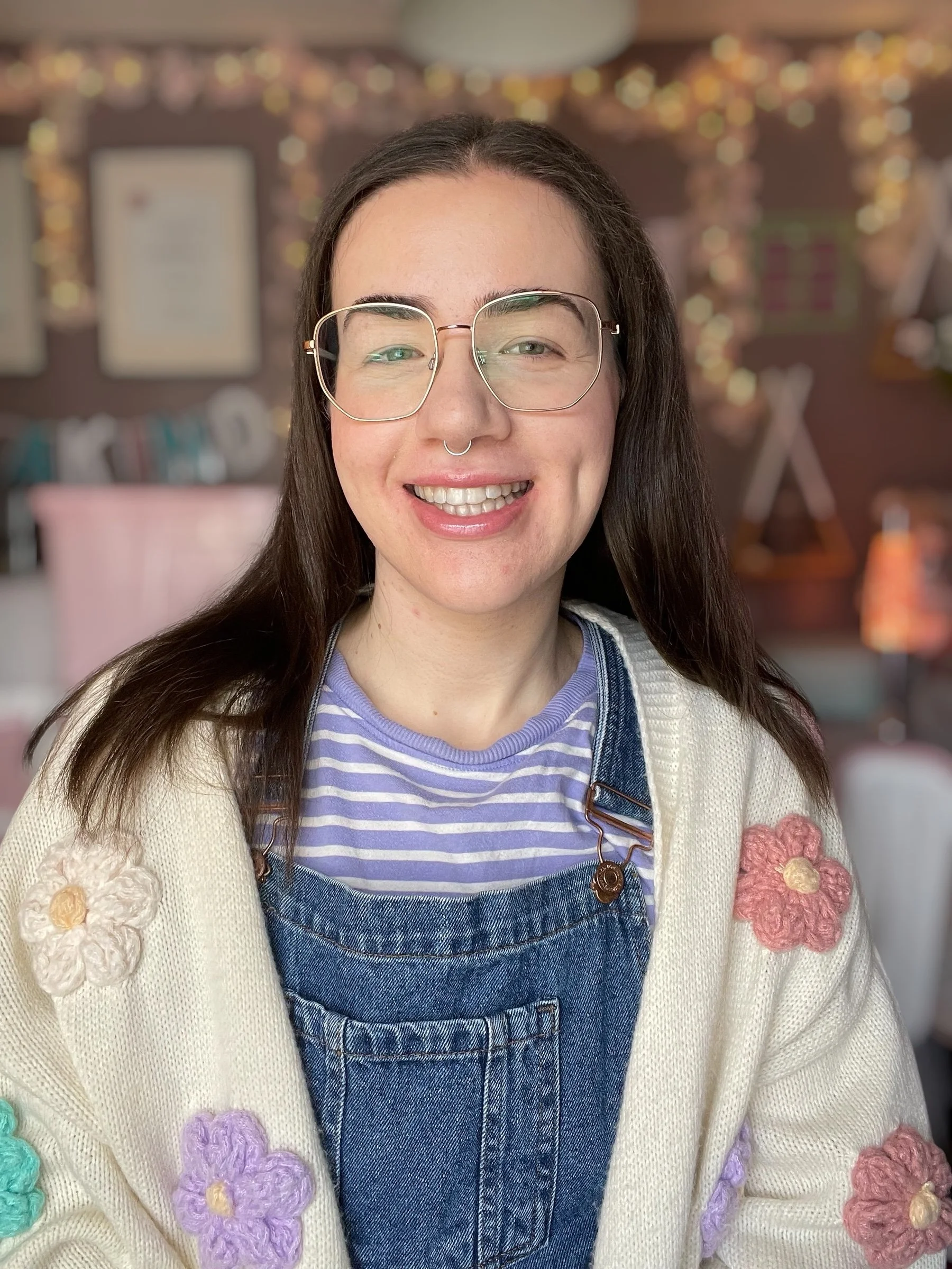 A woman with glasses and a septum piercing smiling, wearing a pastel-colored cardigan with knitted flower embellishments over a striped shirt and denim overalls, with a decorated background of blurred lights and wall art.