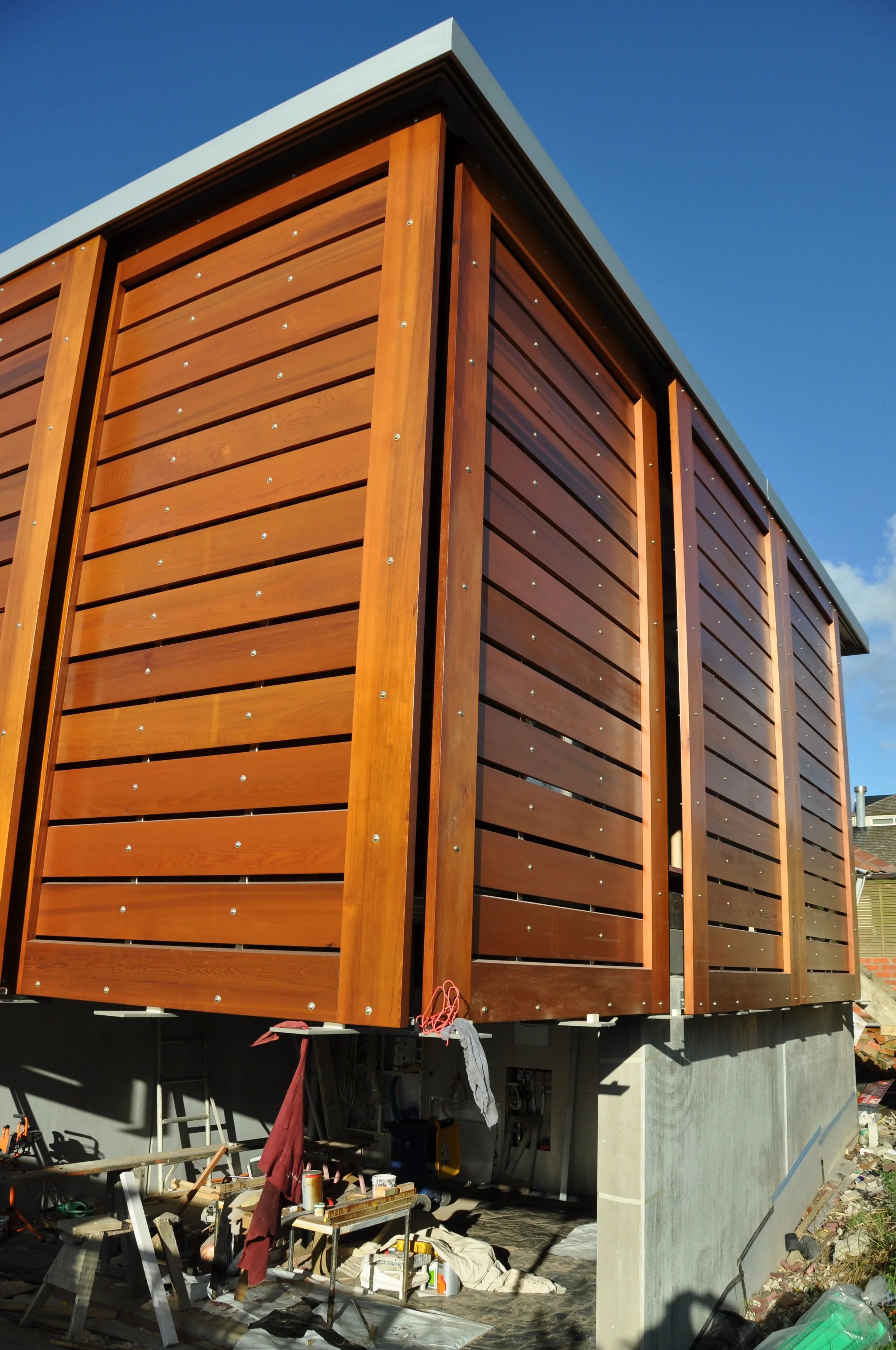 Construction site with a building featuring wooden siding, with construction tools and materials scattered at the base, under a clear blue sky.