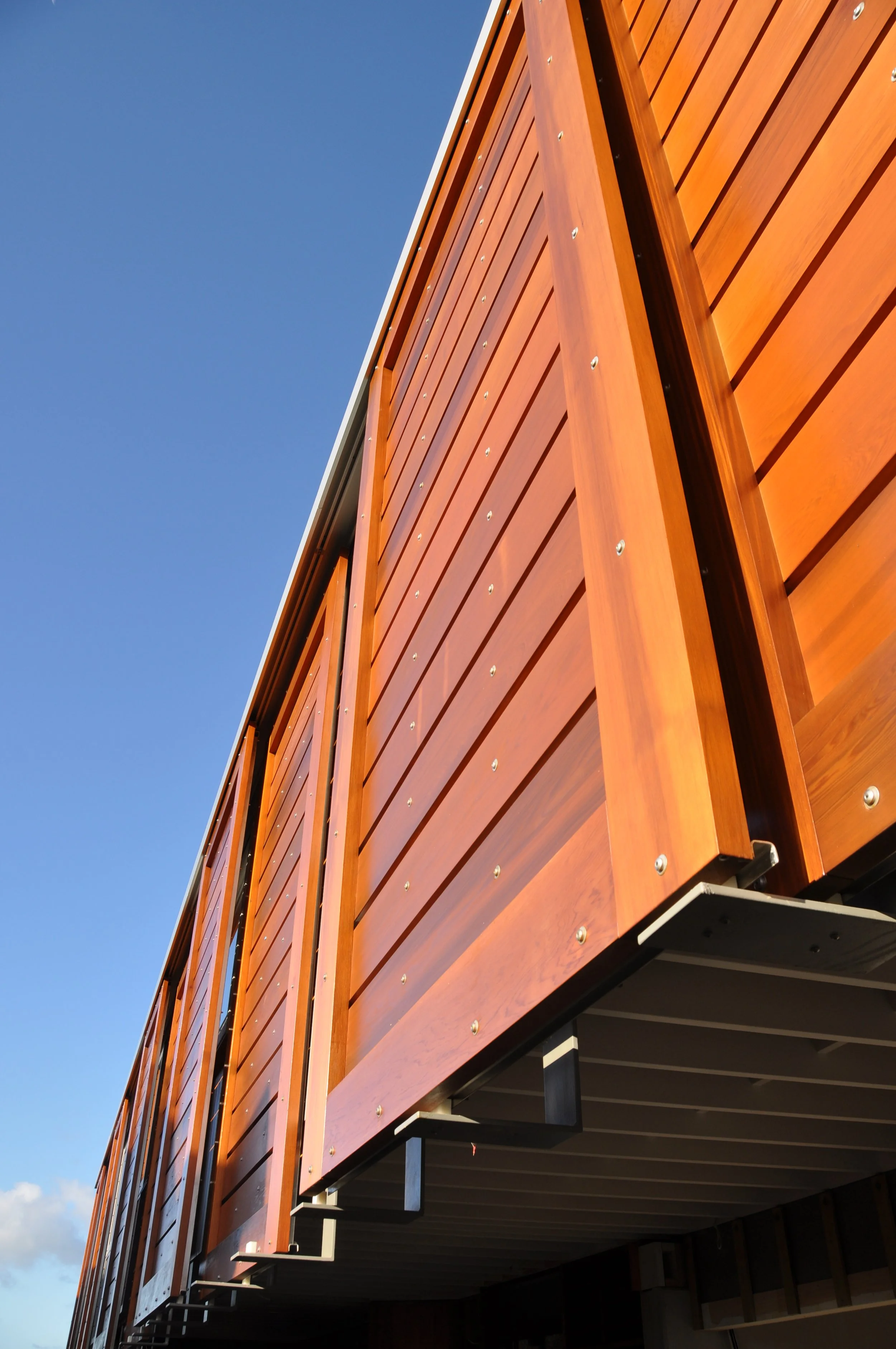 Close-up of a modern building's exterior with wooden panels and metal support beams, against a clear blue sky.