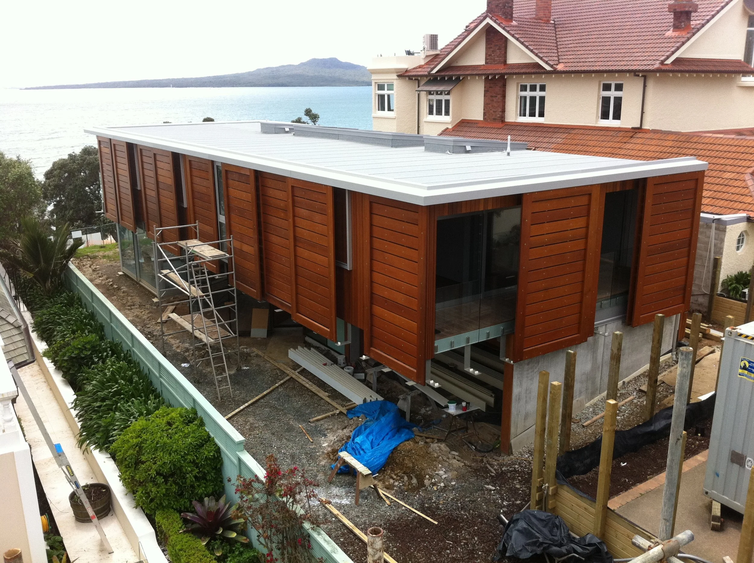 Construction site of a modern house with wooden exterior walls, large glass windows, and a metal roof, overlooking a body of water with mountains in the background.