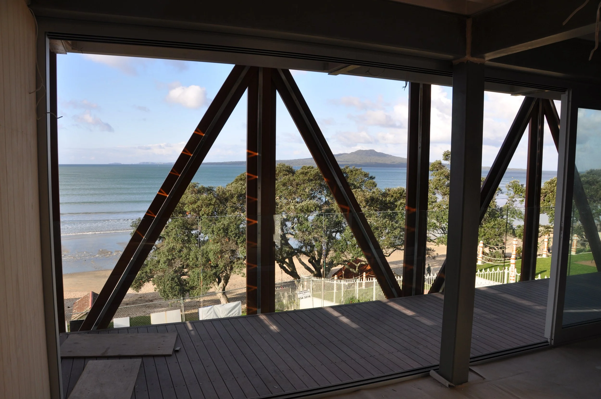 Interior view of a building under construction, showing large windows with a scenic ocean and mountain view outside. The interior has wooden flooring and structural beams.