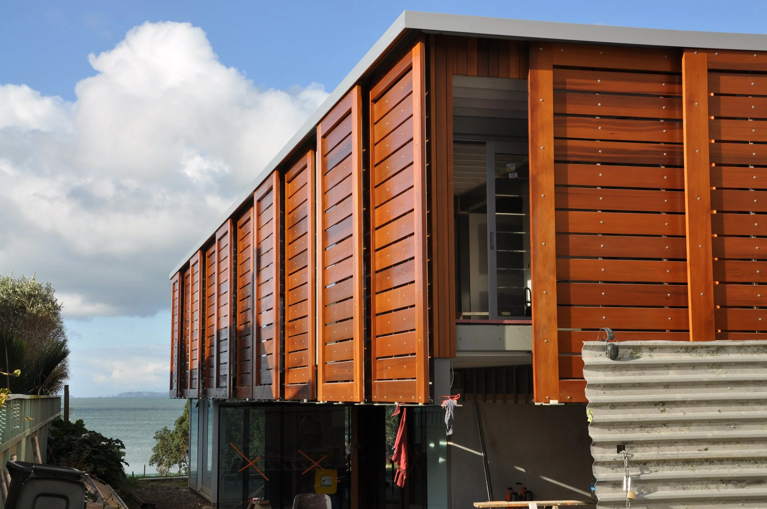 A modern building under construction with wooden exterior panels, large glass windows, and concrete stairs leading to the entrance. The background shows a partly cloudy sky and trees near a water body.