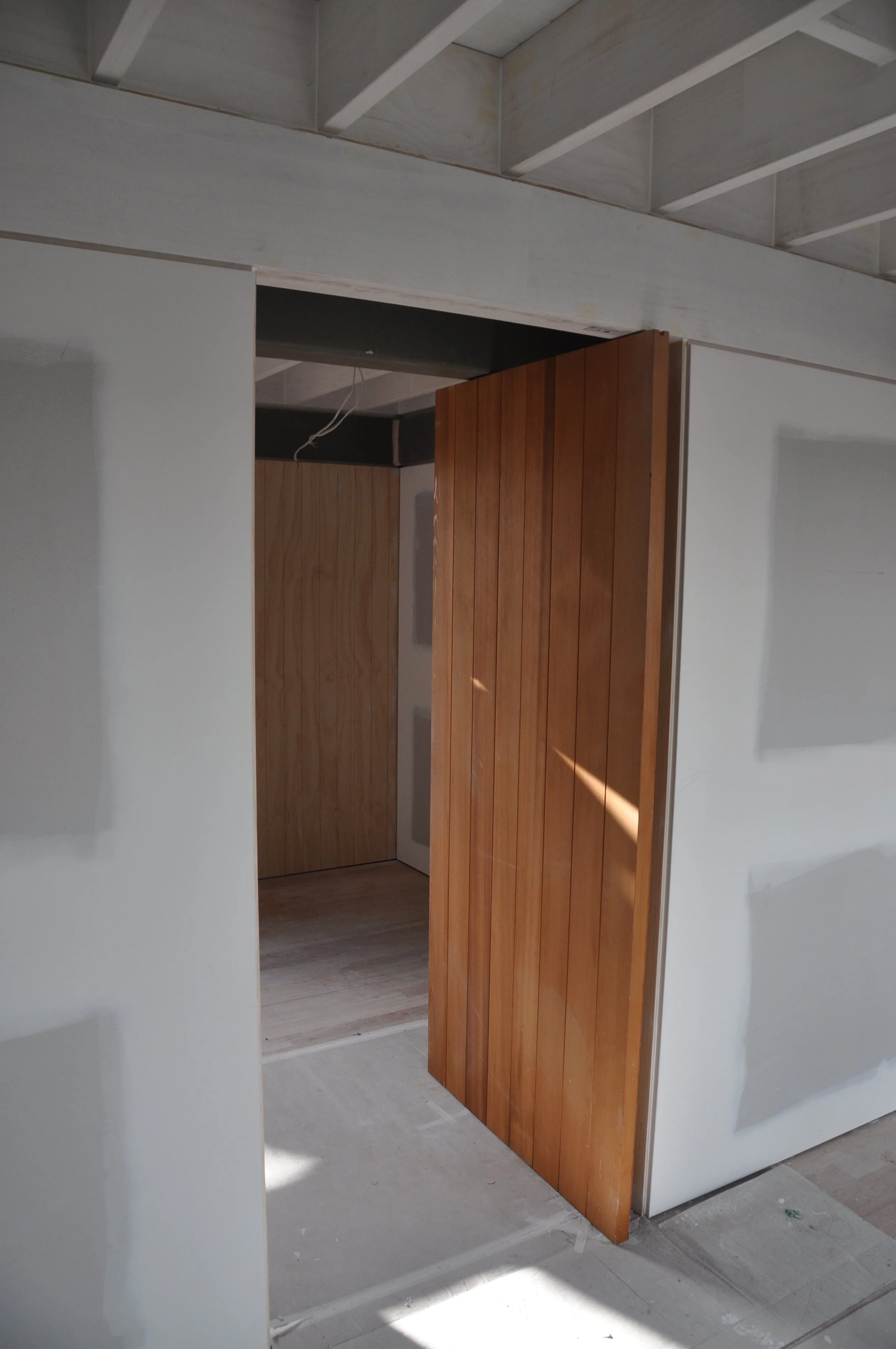Wooden folding door partially opened in a room under construction with unfinished ceiling and white walls.