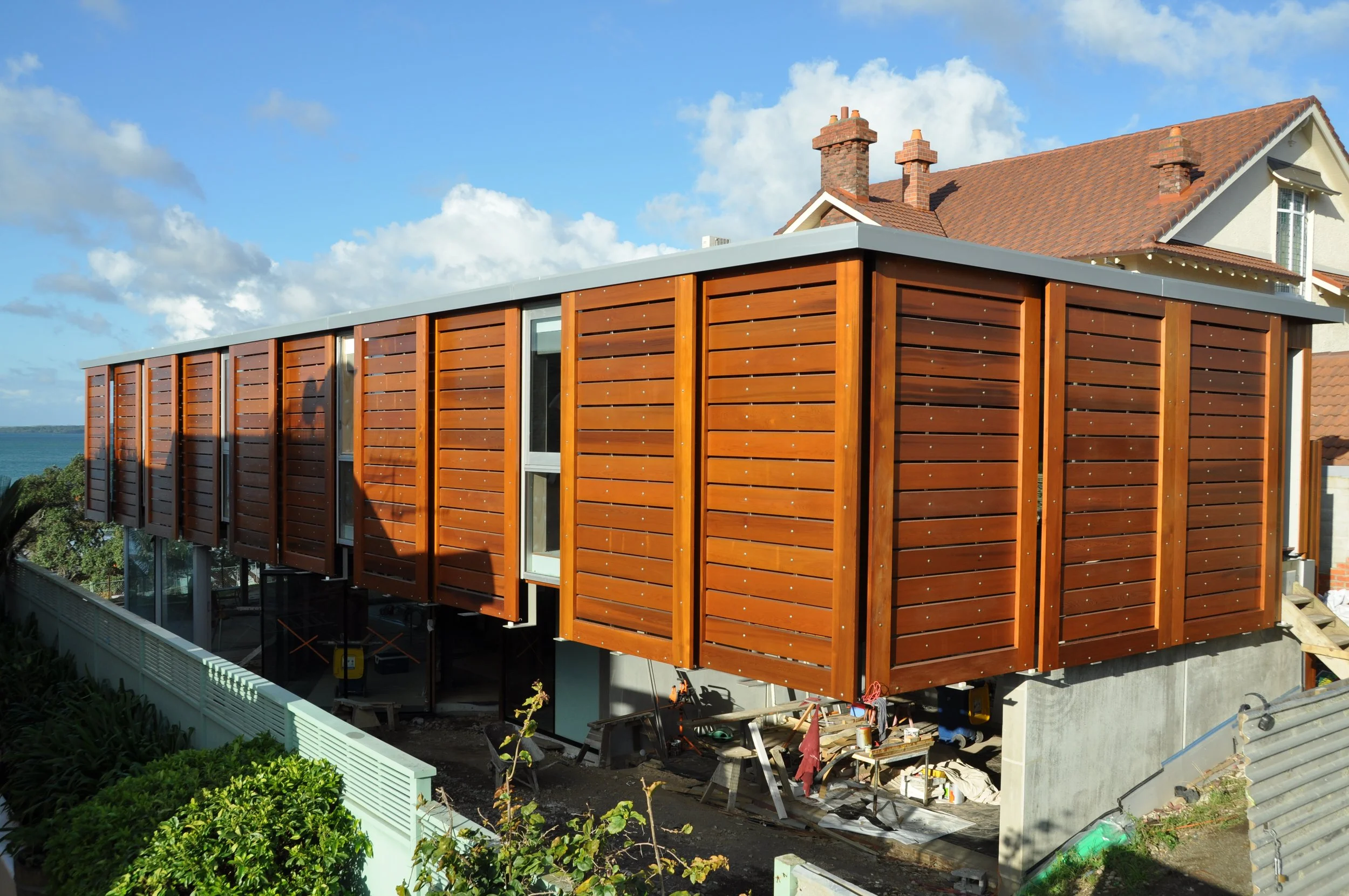 Modern building with wooden siding under construction, with residential house with brick chimneys in the background, blue sky, and some clouds.