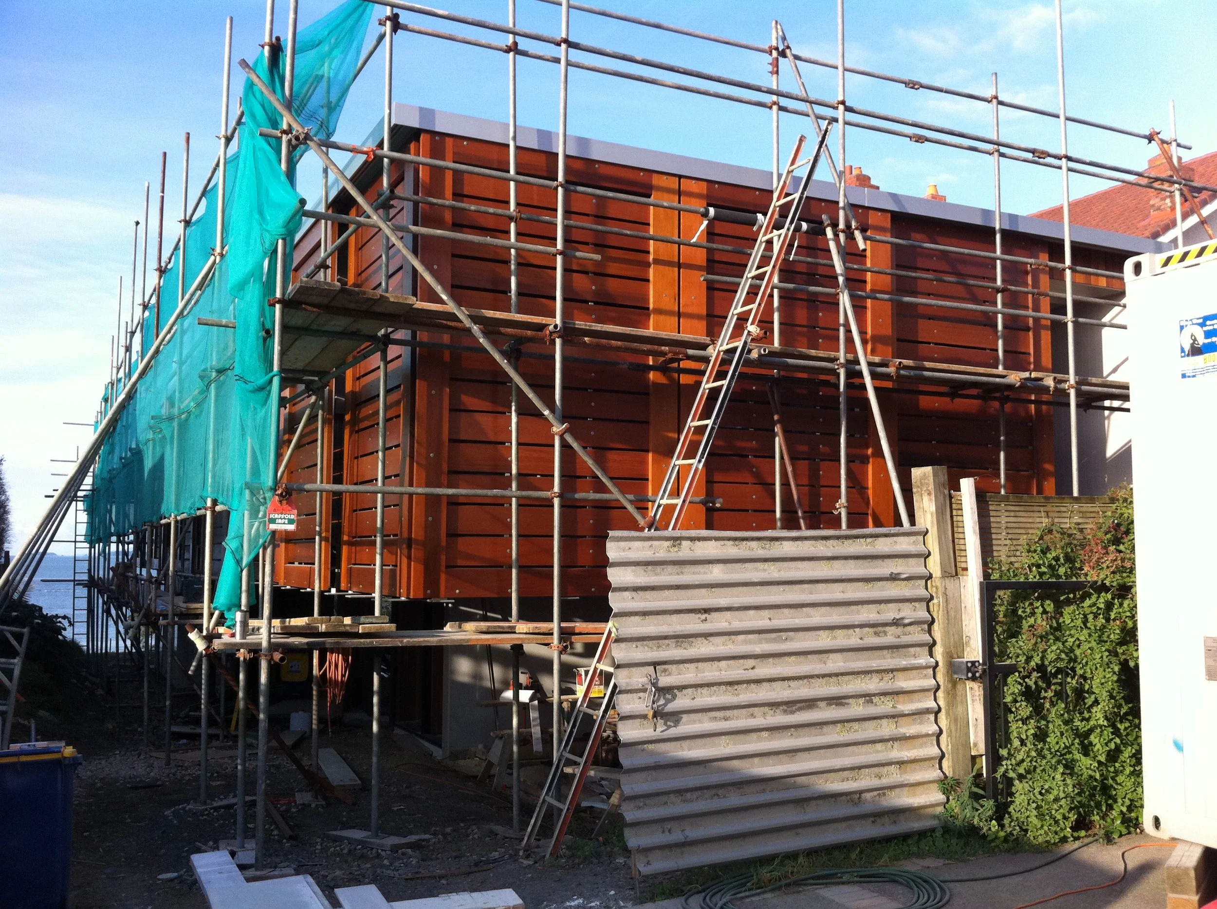 A house under construction with scaffolding surrounding it. The house has a modern design with horizontal wooden panels on the exterior. There are steps leading up to the entrance, and construction materials and equipment are visible around the site.