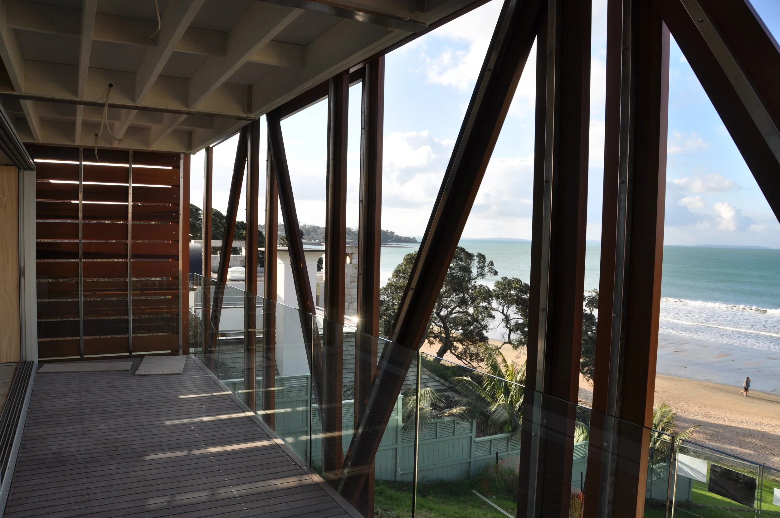 Interior view of a modern building with large glass windows and wooden beams, overlooking the ocean and beach with trees and a person walking.