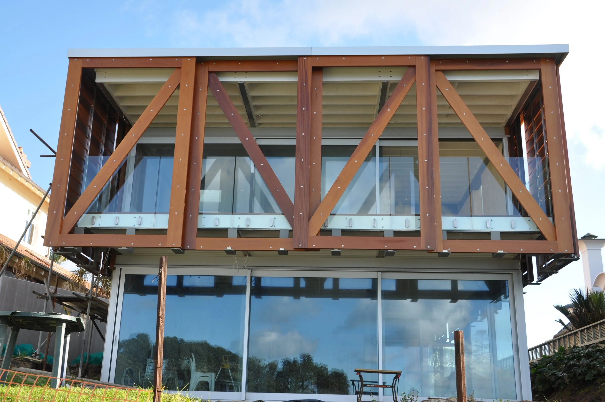 Front view of a modern house under construction, featuring wooden framing on an upper balcony, glass sliding doors on the lower floor, and a clear blue sky.