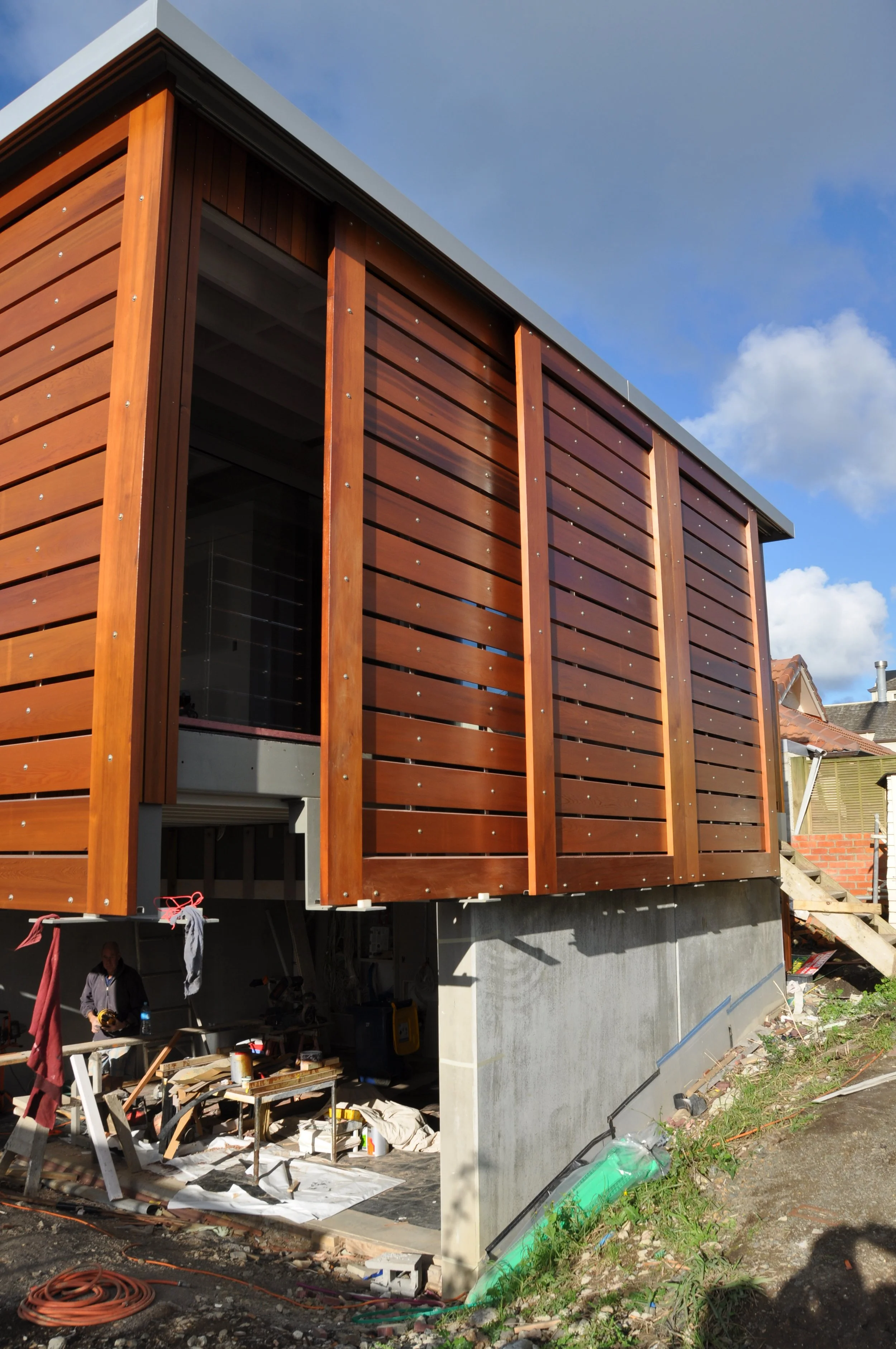 Construction site of a modern building with wooden exterior siding, partial view of a person working at a table, and building materials scattered on the ground.