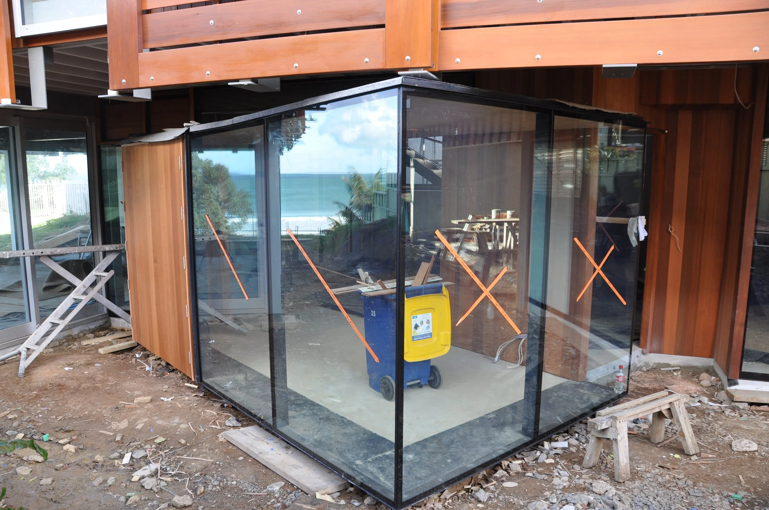 Construction site with a glass-enclosed structure on a concrete slab, surrounded by wooden building materials, equipment, and a trash bin, overlooking the ocean in the background.