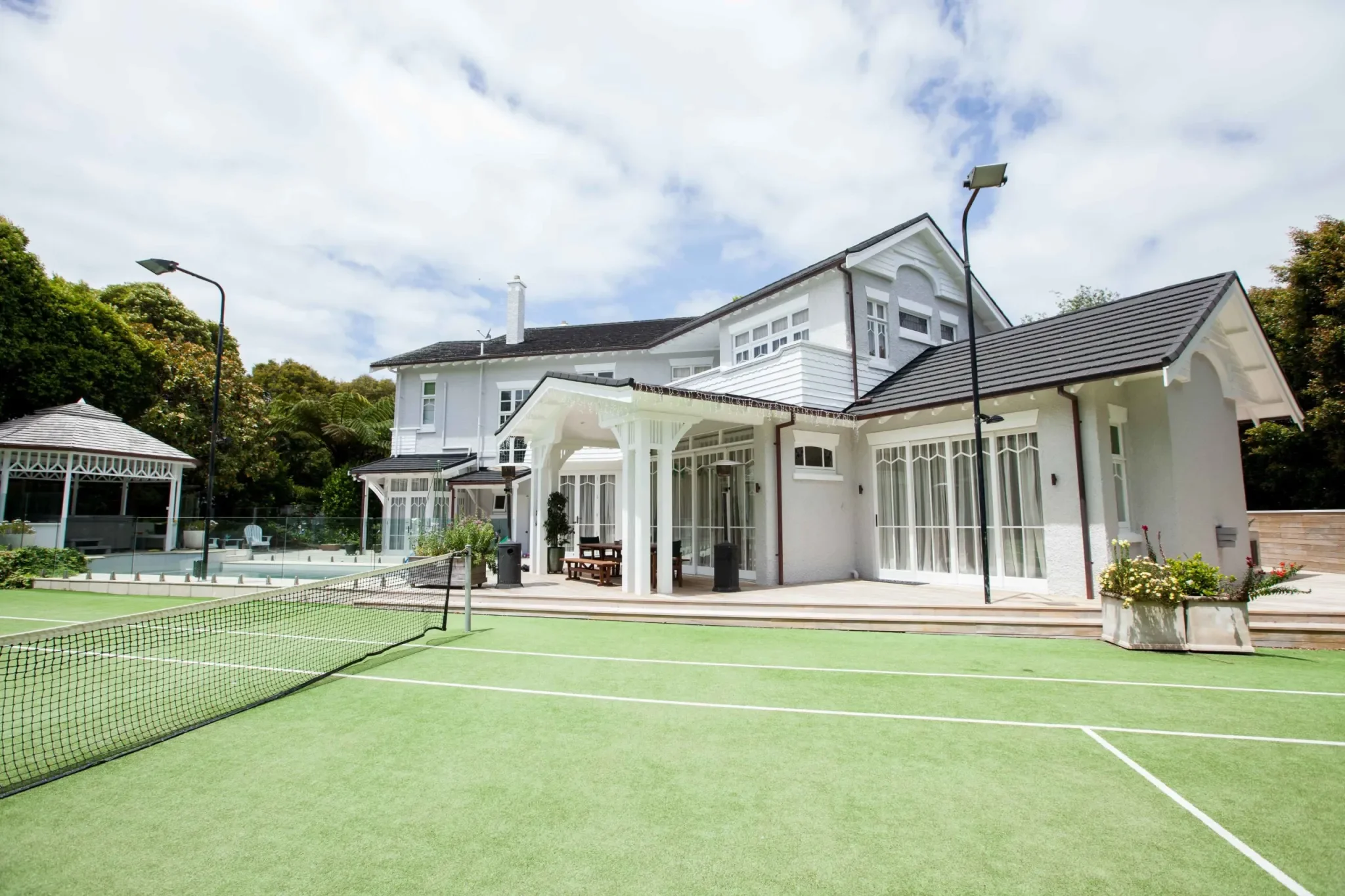 A backyard tennis court with a large white house in the background, surrounded by trees and a sky with clouds.