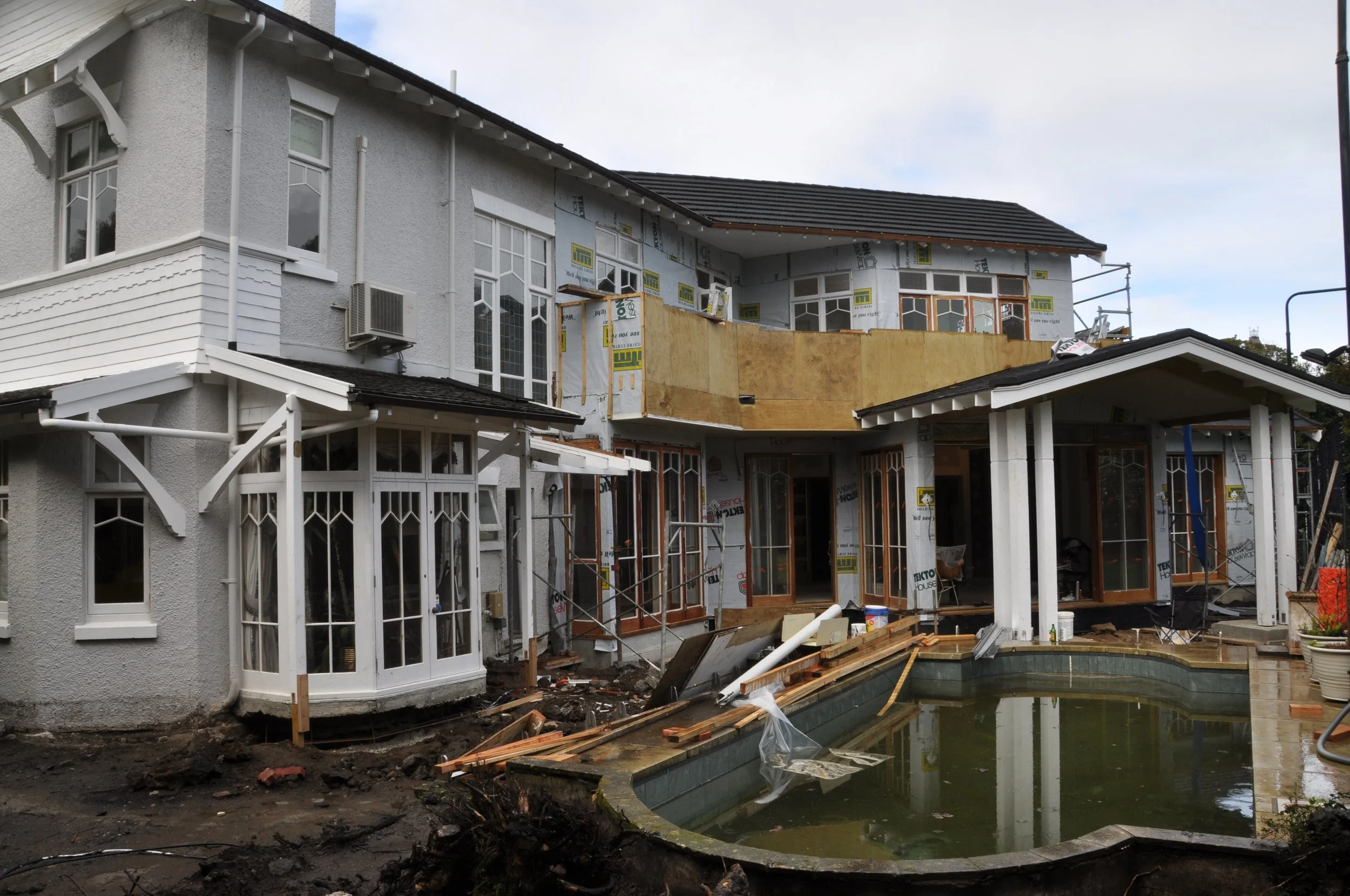 A house under construction with scaffolding, an unfinished balcony, and a rectangular swimming pool in the backyard.