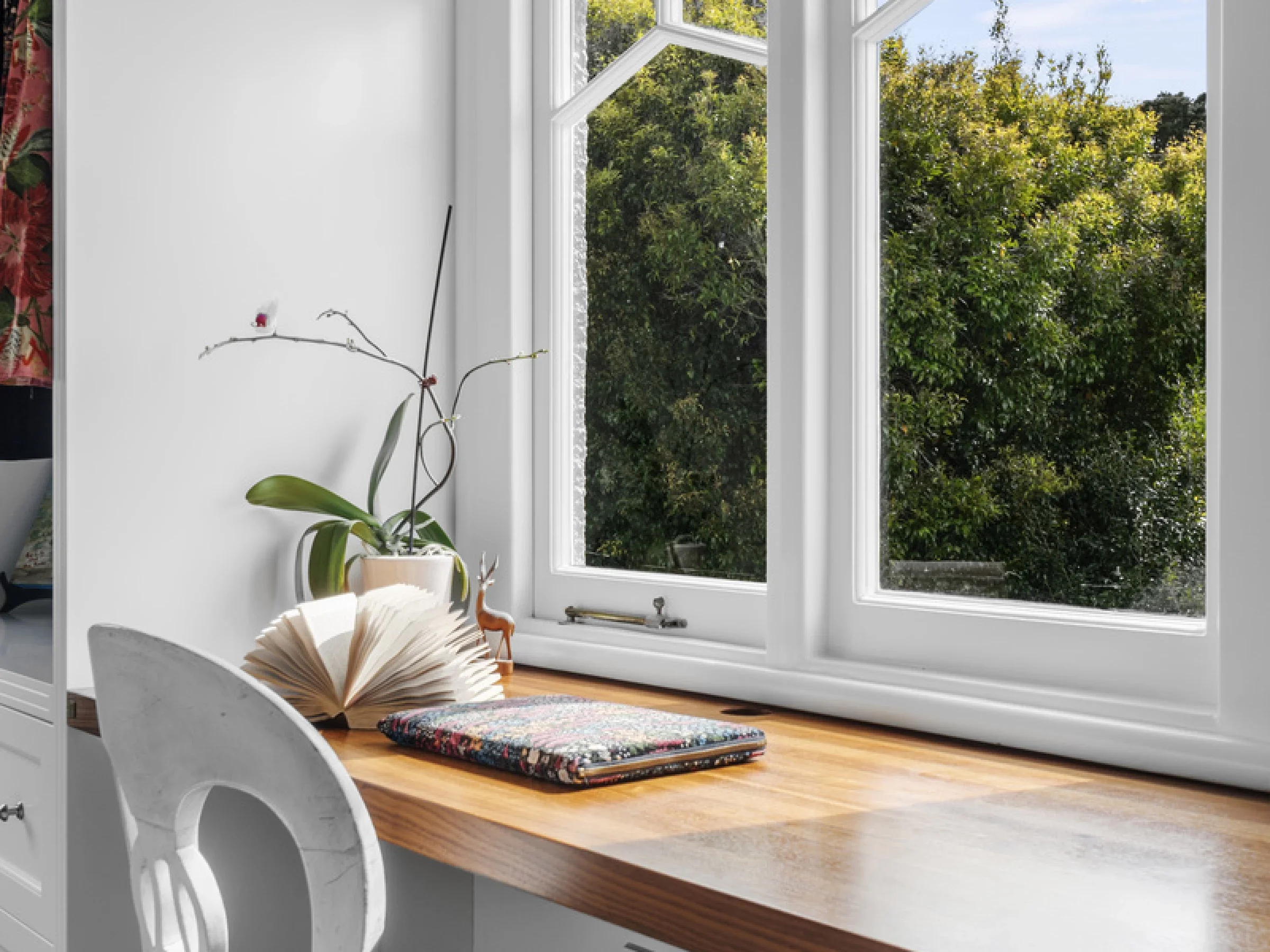 A wooden desk with an open book, pink and multicolored pillows, and a crocheted mat in front of a large window with white frame showing green foliage outside.