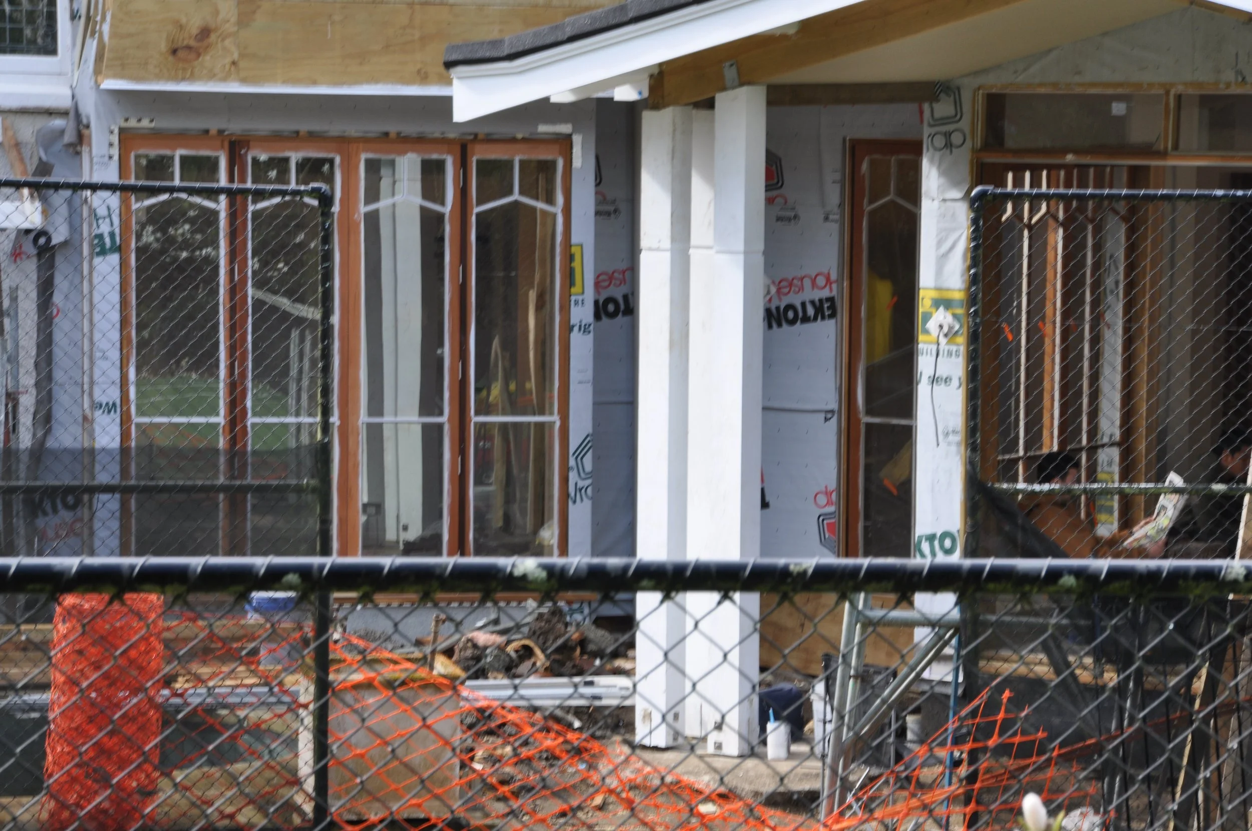 Construction site with unfinished building structure, windows, and a chain-link fence in the foreground, surrounded by construction materials and orange safety netting.