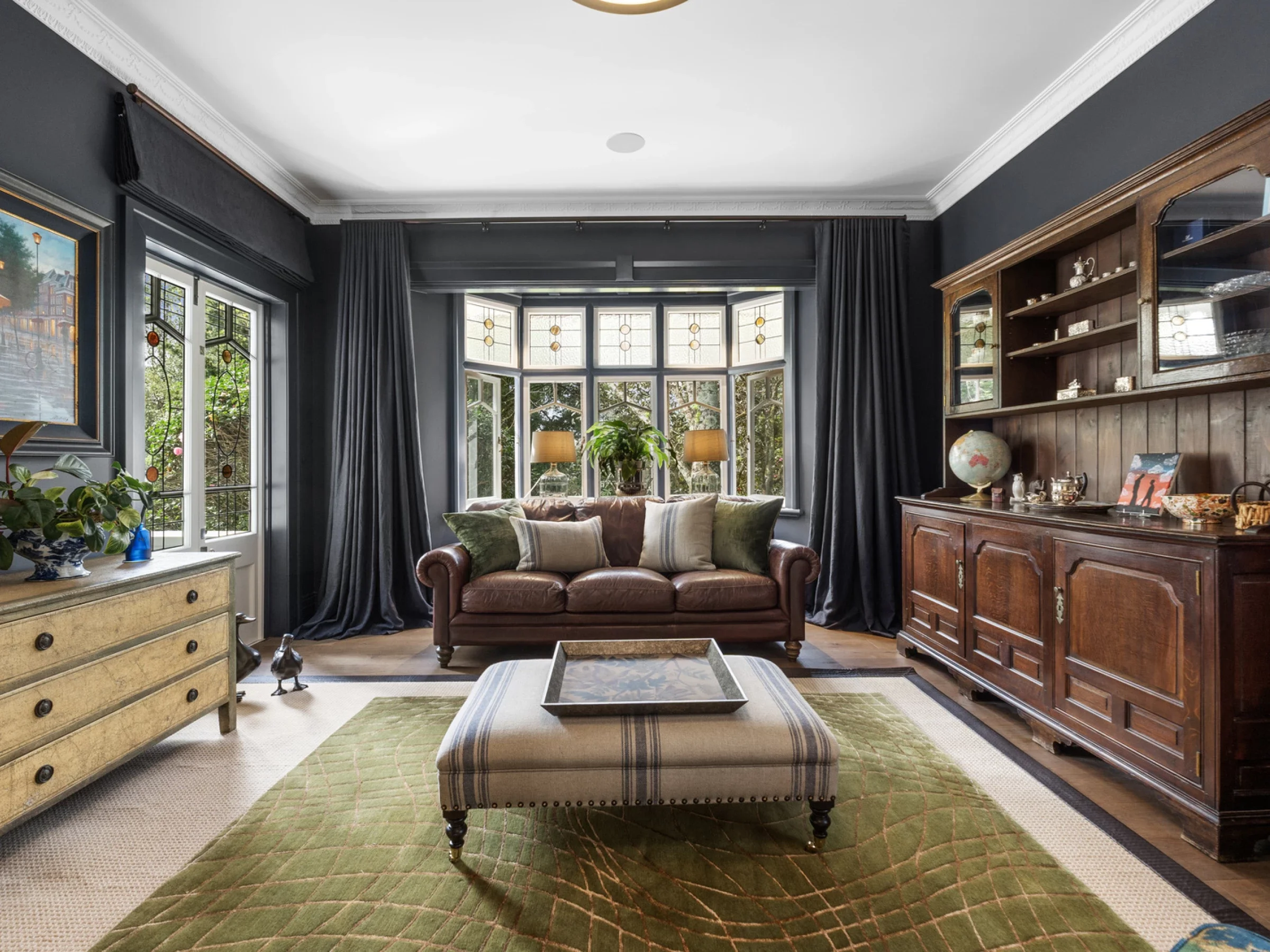 A cozy living room with dark walls, large bay windows, and vintage furniture. There is a leather sofa, a green rug, a wooden sideboard with a globe and decorative items, and a glass coffee table. Houseplants and artwork adorn the space.