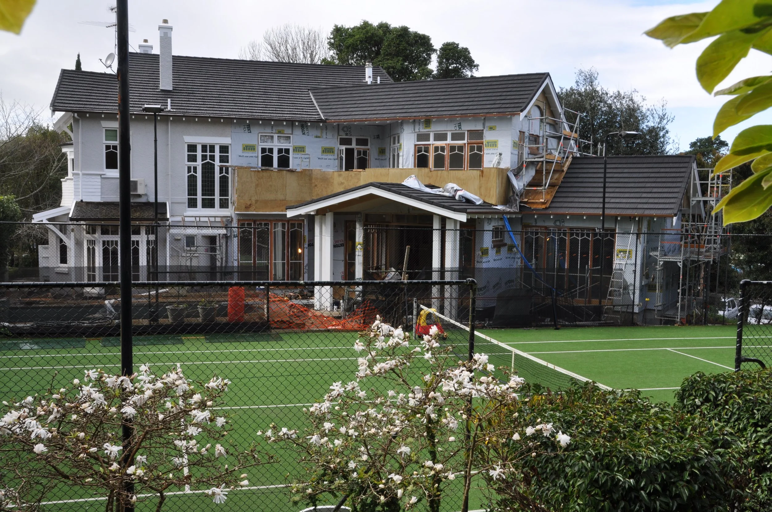 A house with scaffolding and construction materials on the upper floor, surrounded by a chain-link fence, with a green tennis court and flowering bushes in the foreground.