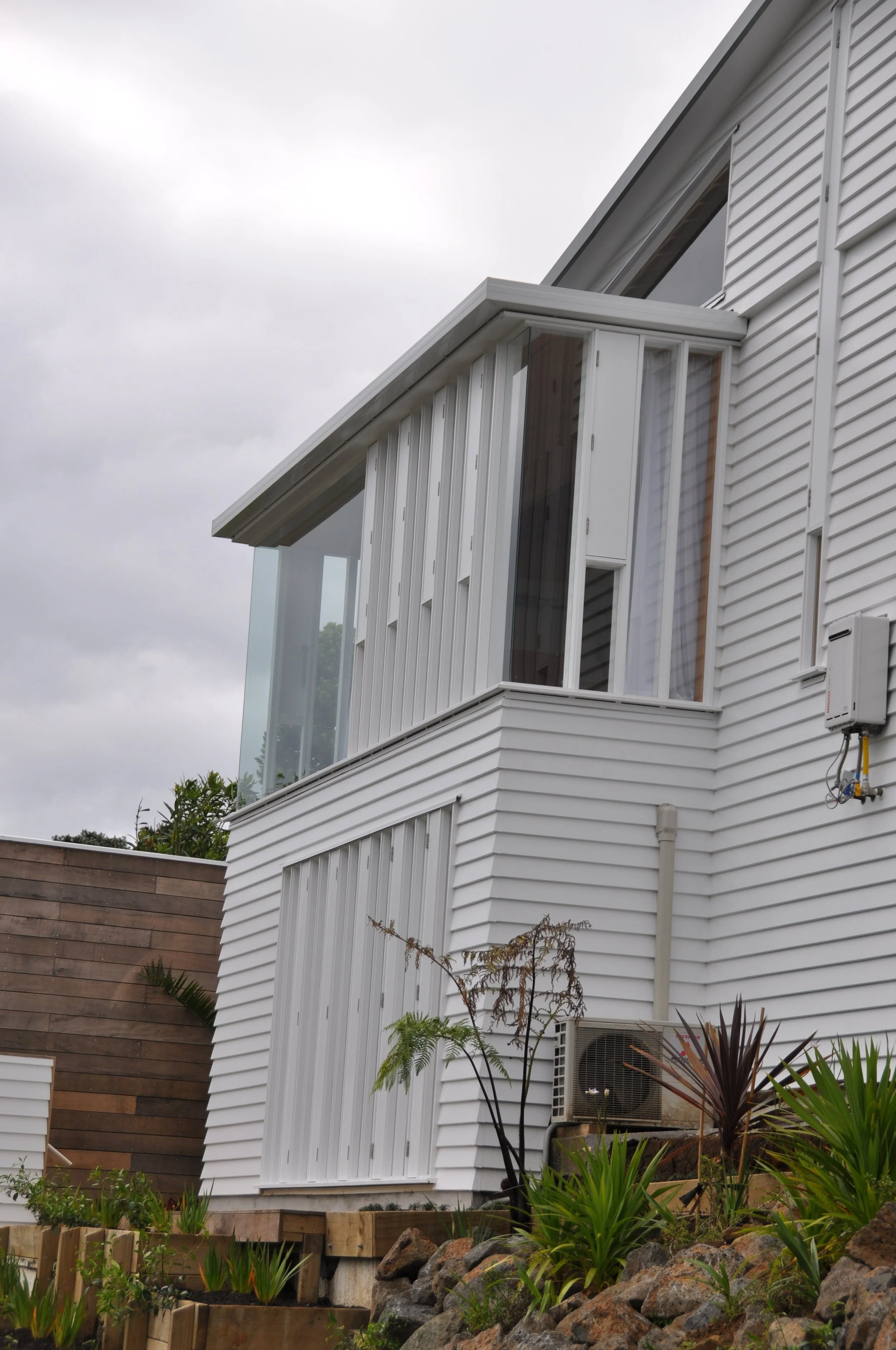 The image shows a modern white house with large windows and a small balcony, set against a cloudy sky. There is a wooden fence to the left and a garden with plants, rocks, and a small tree in the foreground.