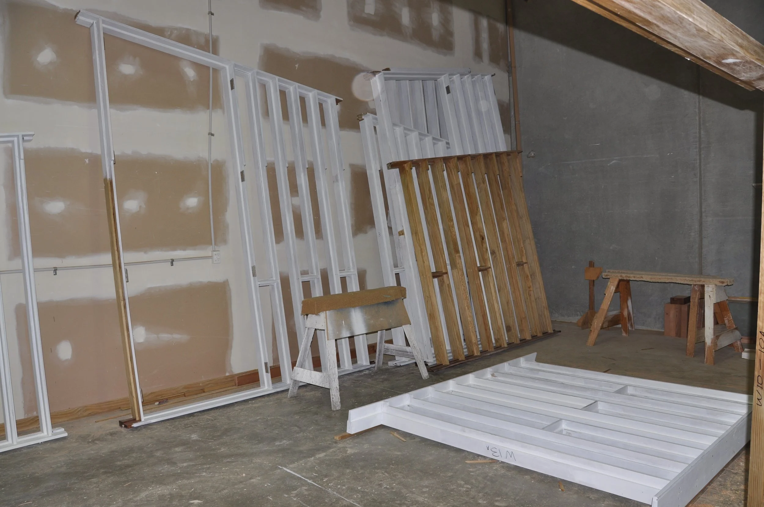 Various white and wooden window frames and wooden slats stored in a workshop against a wall with unfinished drywall. A wooden workbench and chair are also visible.