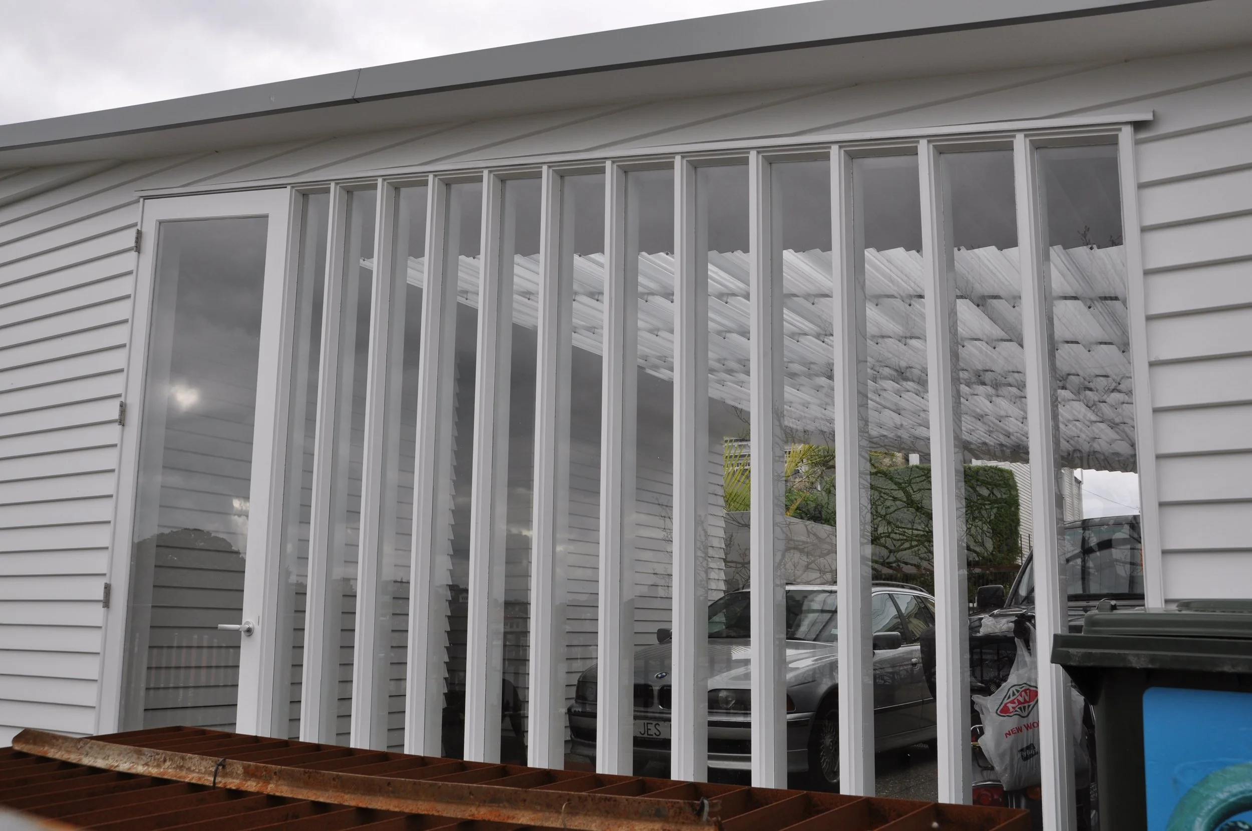 Side view of a white house with horizontal siding and a large window with vertical bars, garage roof and parked cars visible through the window.