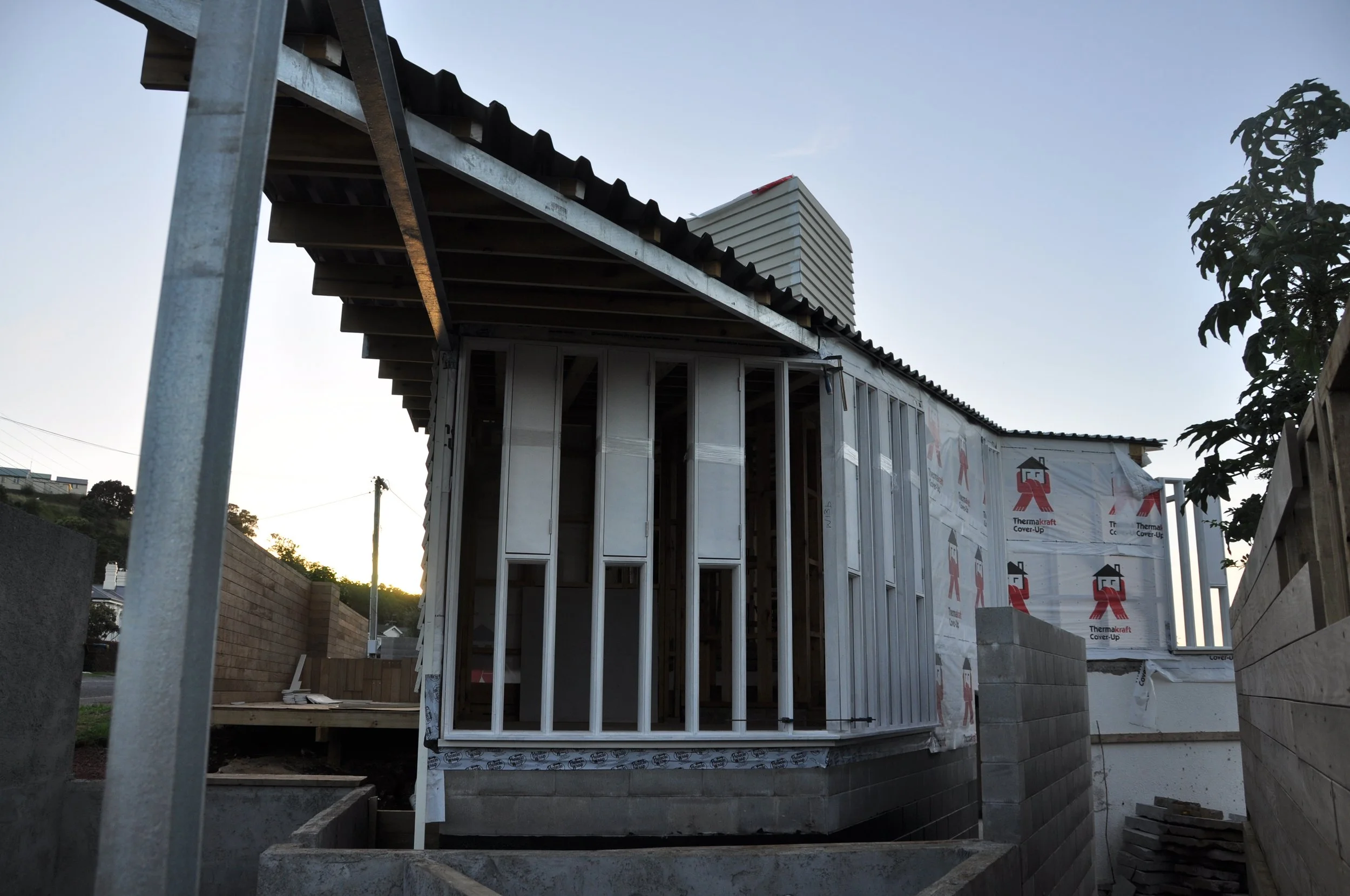Construction site with a building frame and insulation wrap, showing part of a house under construction during sunset with a tree on right side.
