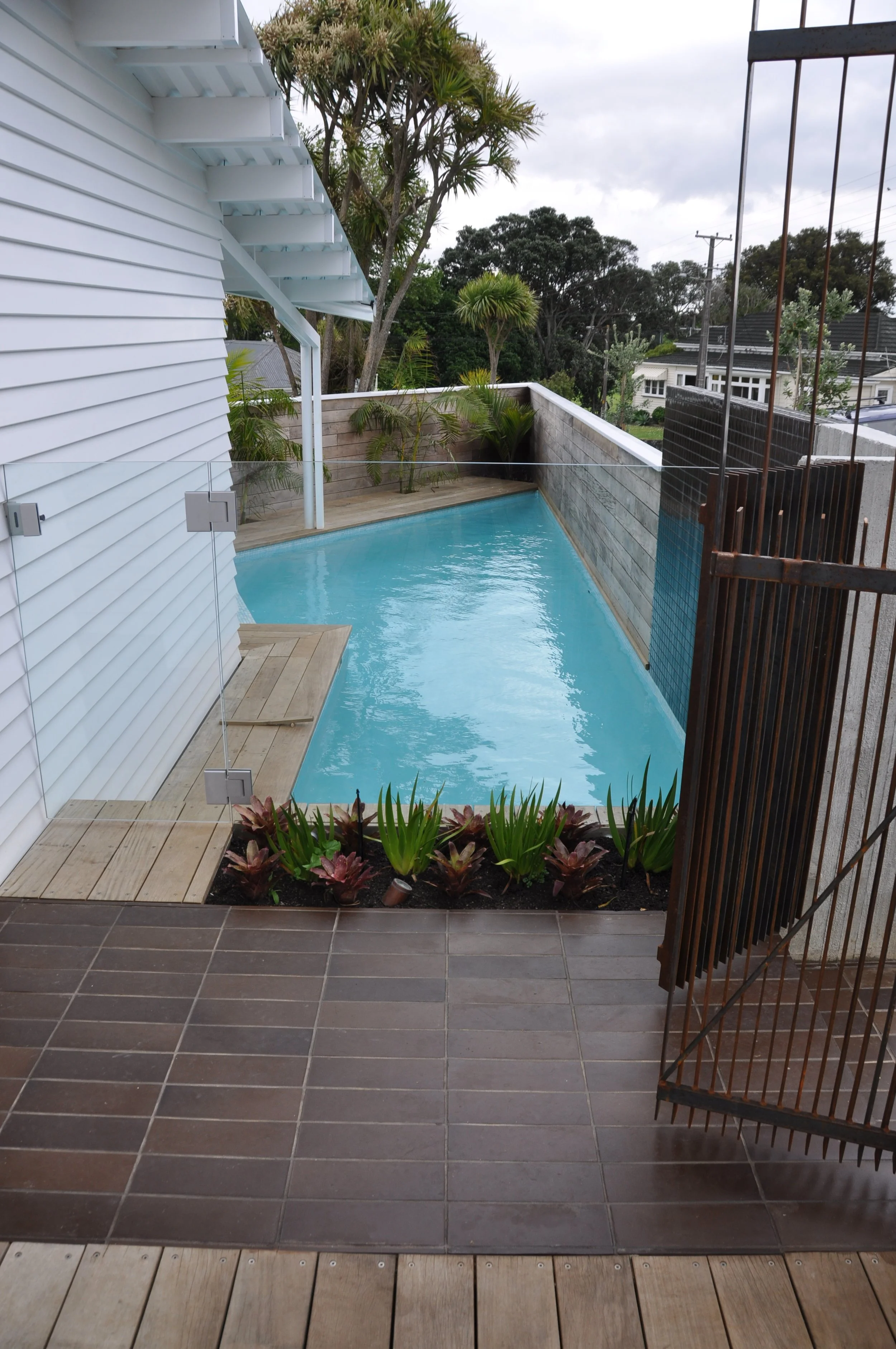 View of a small private backyard in a suburban neighborhood, showing a swimming pool, surrounding wooden deck, plants, and neighboring houses with trees overcast sky.