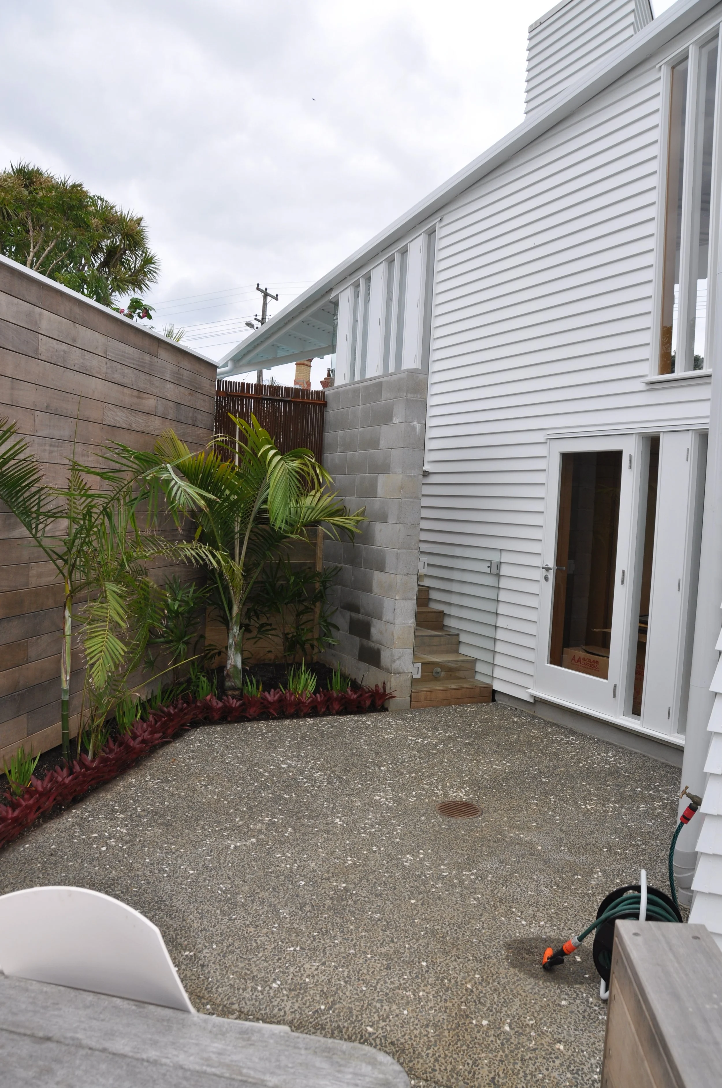 Backyard patio with small garden bed featuring various plants, a wooden fence, a white house with sliding glass doors, and outdoor equipment like a garden hose and a garden light.