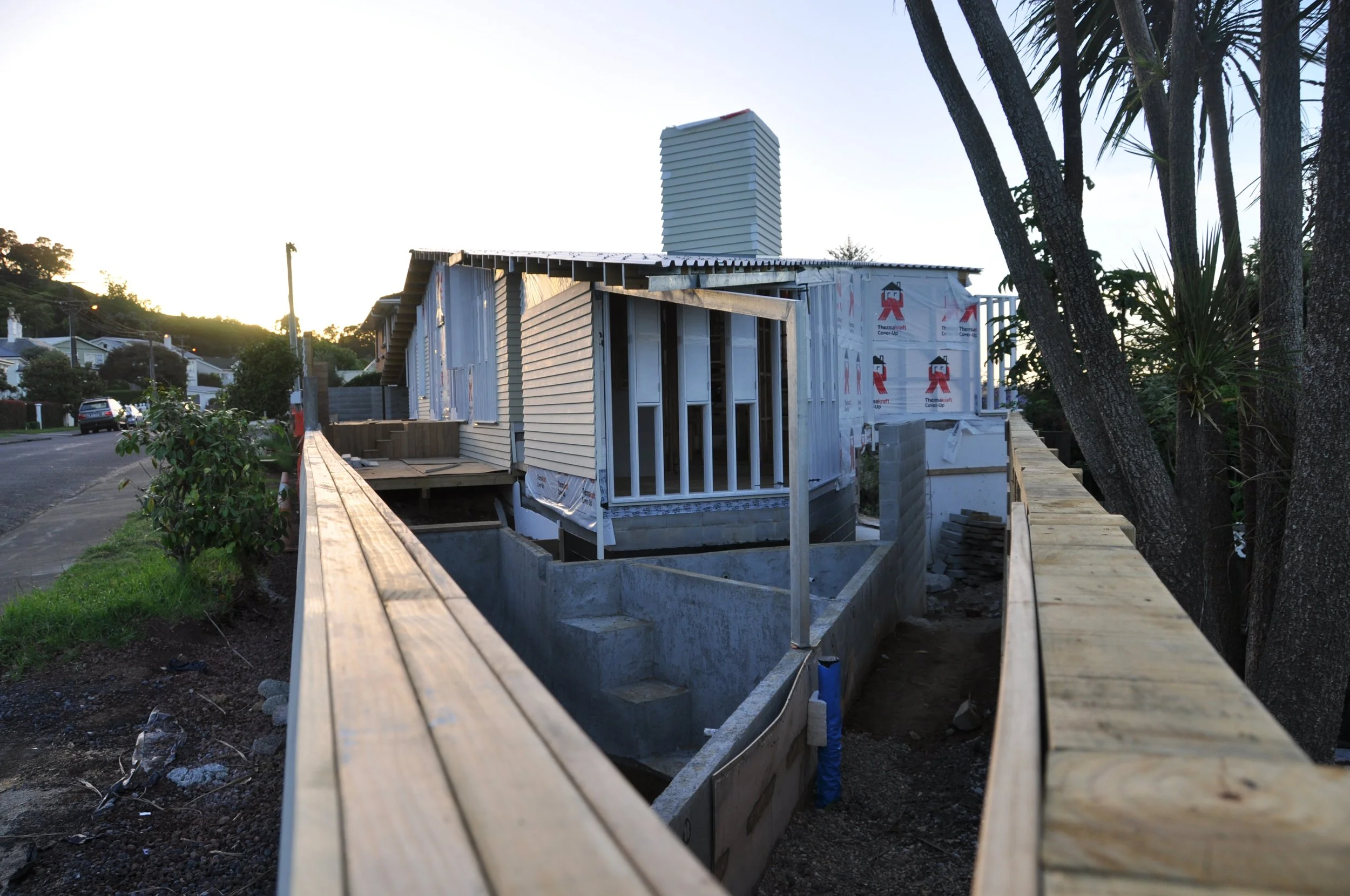 Construction of a house with wooden framing on a suburban street, trees on the right, and a clear sky at sunset.