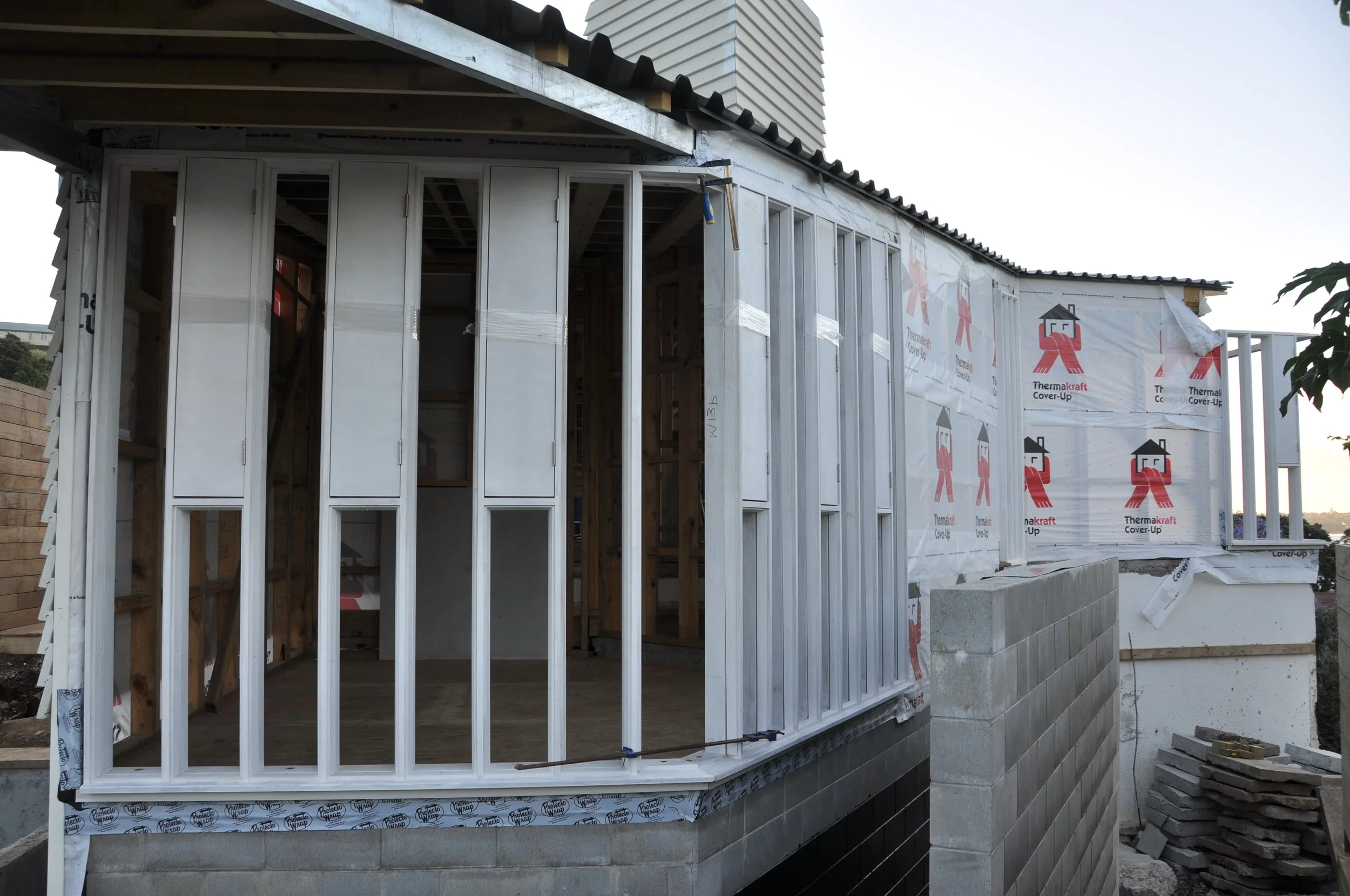 Construction site of a house, with a partially built wall, wooden framework, and weather-resistant house wrap labeled 'Thermakraft Cover-Up'.
