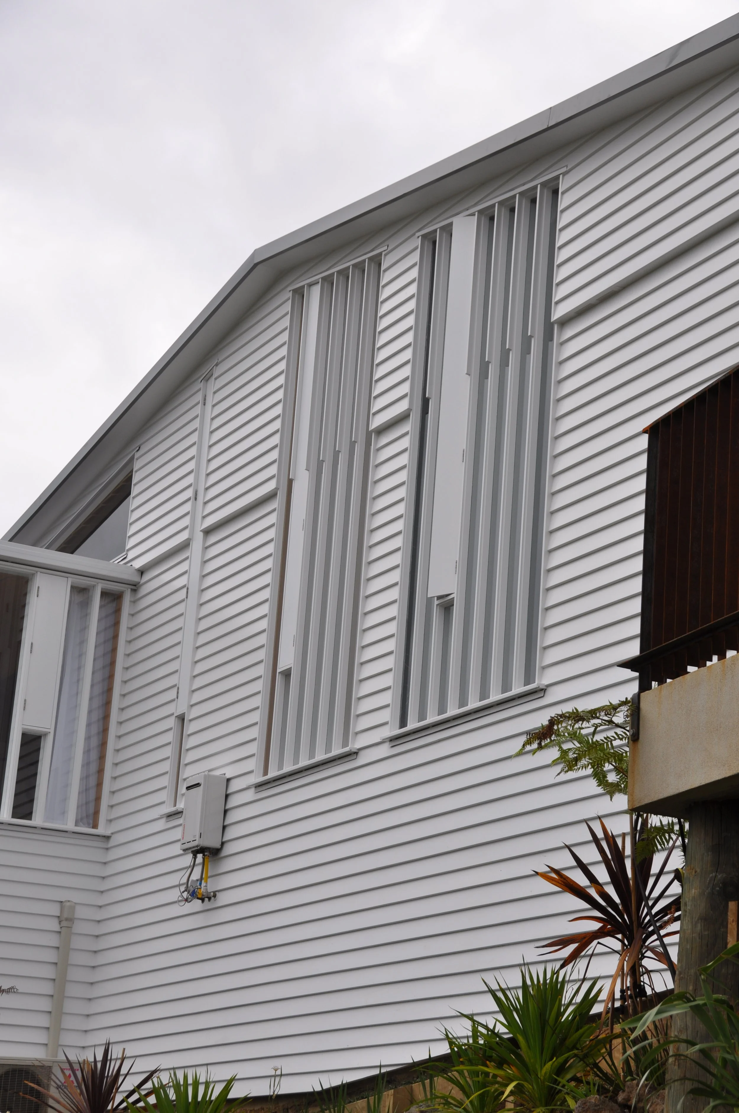 Close-up view of the exterior of a modern white house with horizontal siding, tall narrow windows, and some lush plants near the base.