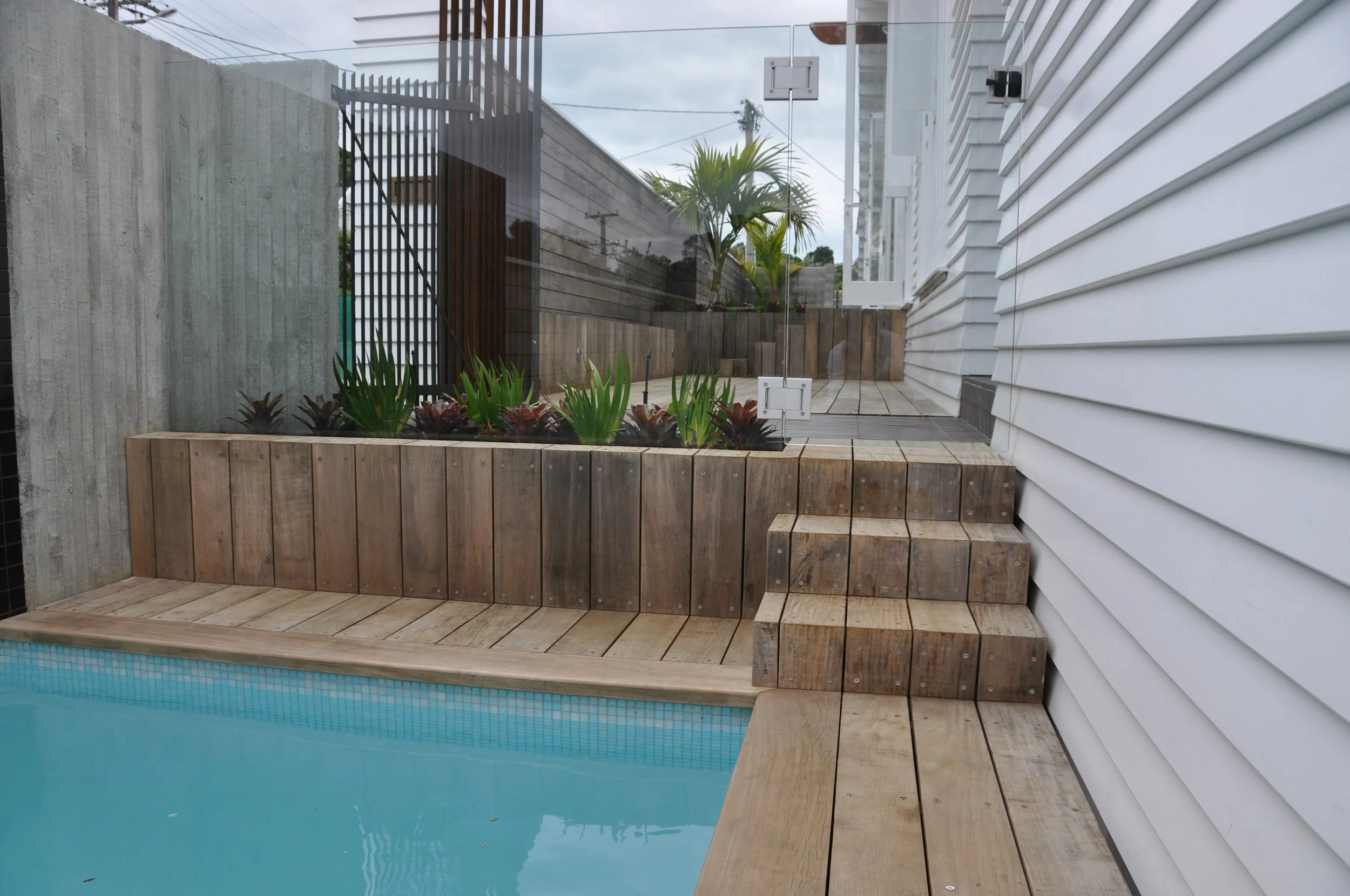 Backyard pool area with wooden decking, a glass fence, and a white house siding. There are some plants inside a wooden planter box and a palm tree outside the fence. Overcast sky in the background.