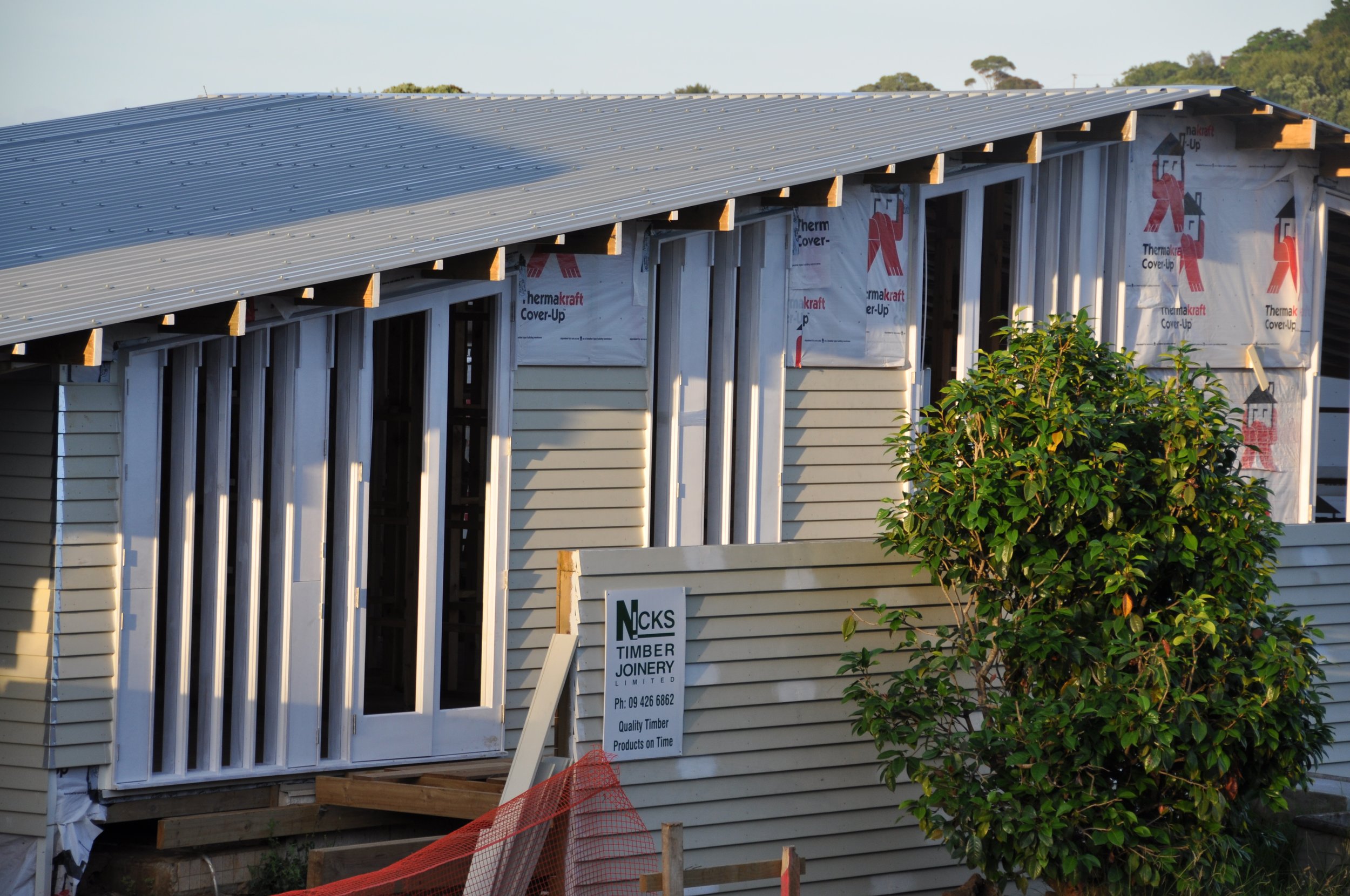 A house under construction with a metal roof, partially installed window frames, and a white siding exterior. There is a green bush in the foreground and a sign with contact information for a timber and joinery company.