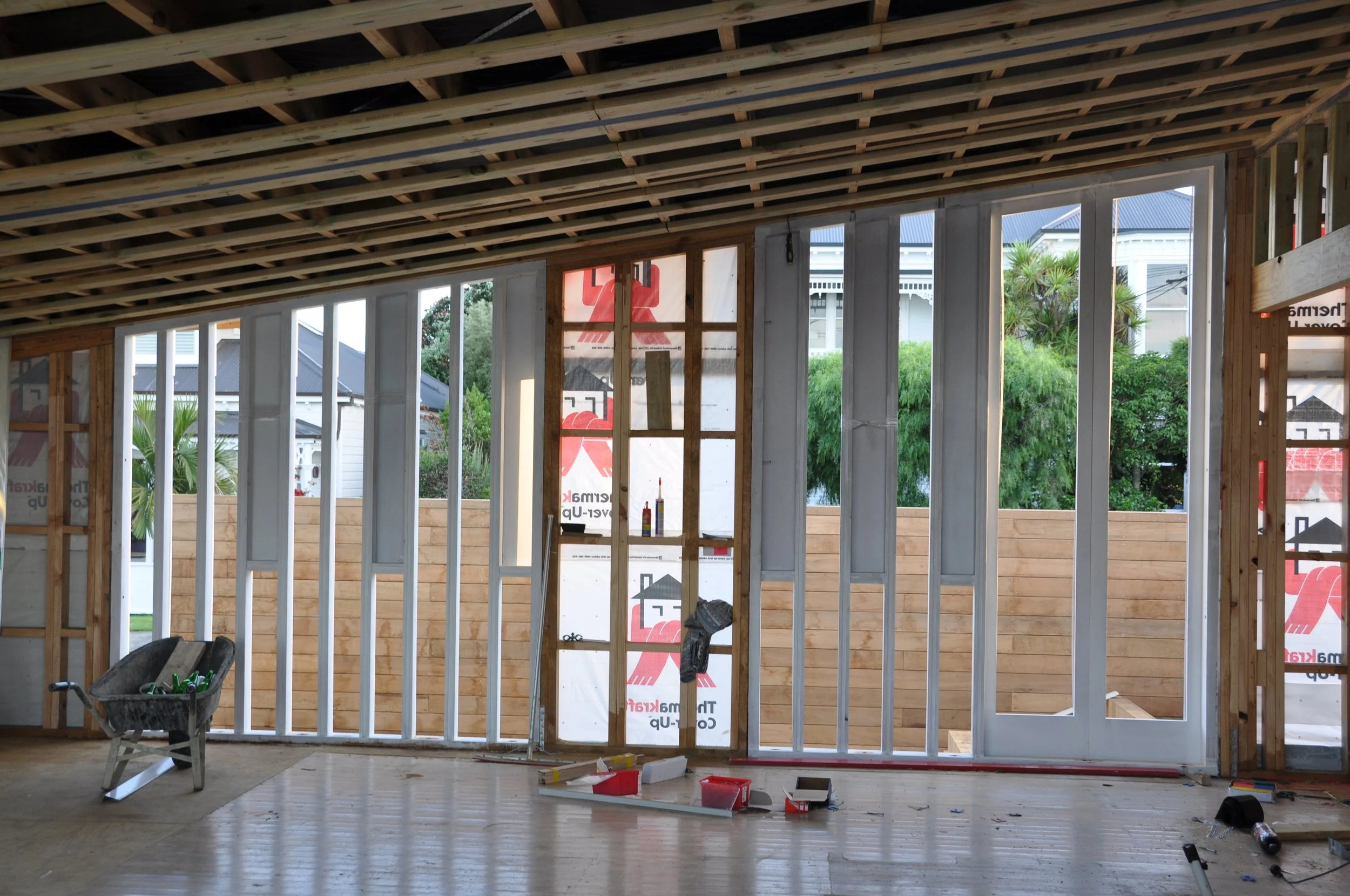 Interior of a house under construction with wooden framing, tools, and materials scattered on the floor, and a view of the outdoors through unfinished wall openings.