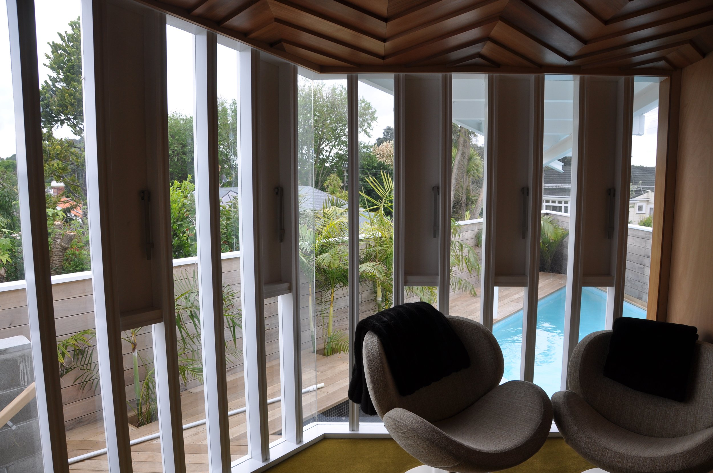 Interior view of a room with large glass windows showing a backyard with trees and a pool, featuring a wooden ceiling and a lounge chair.