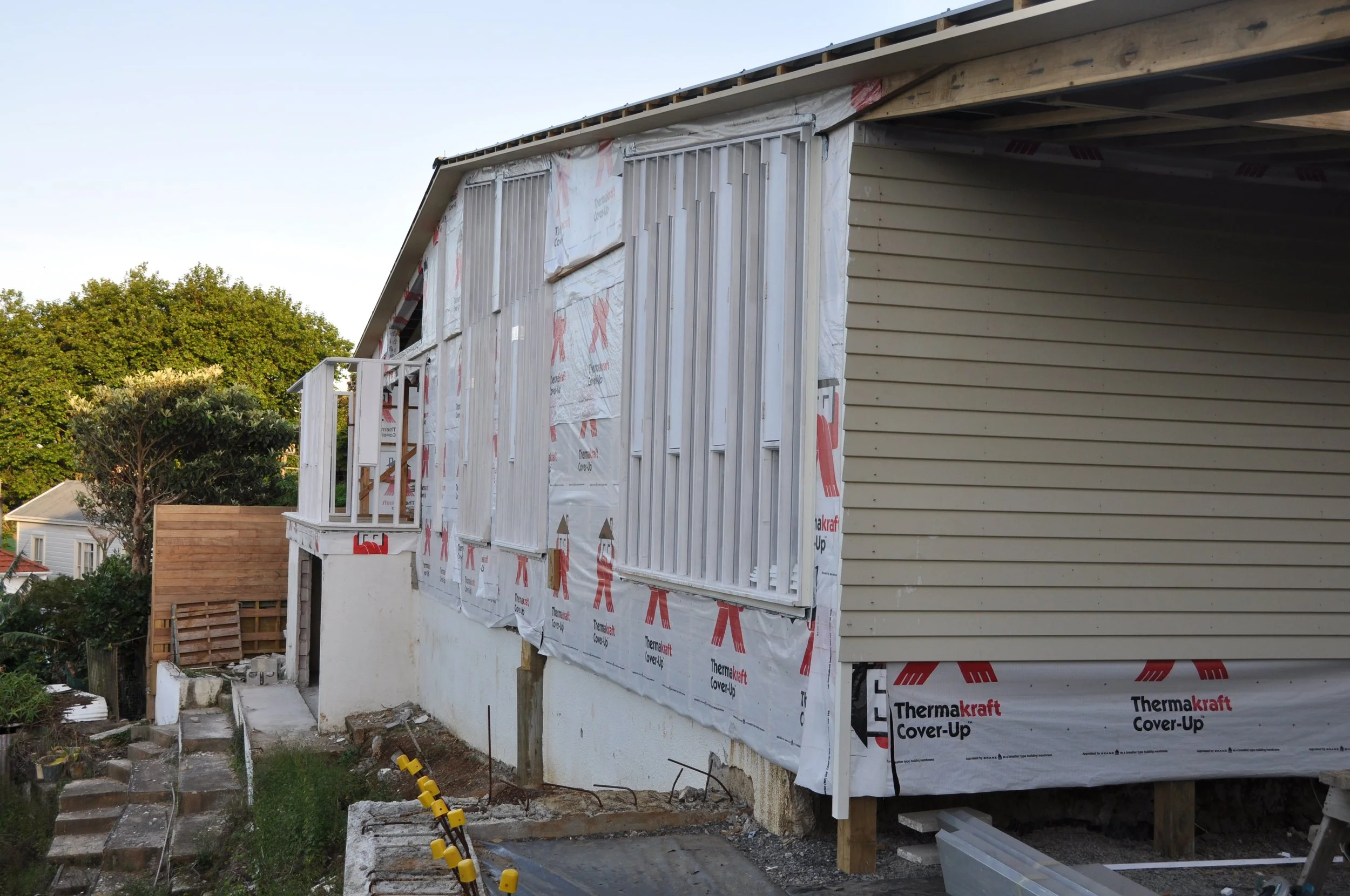 A house under construction with siding partially installed, showing insulation and framing. There are trees and neighboring houses in the background.