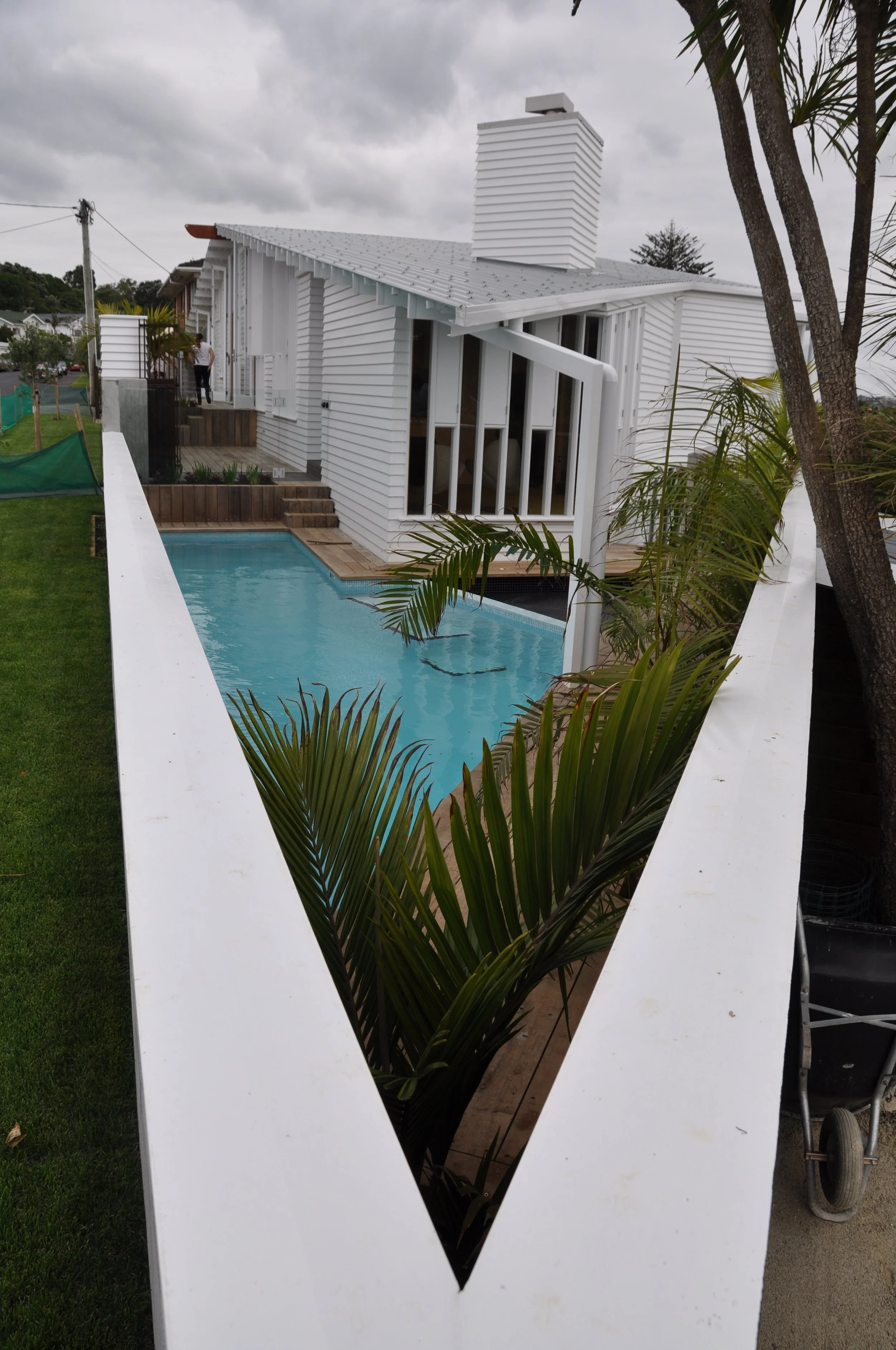 View of a modern white house with a swimming pool, plants, and cloudy sky in the background. The photo is taken through a white fence with a heart-shaped opening.