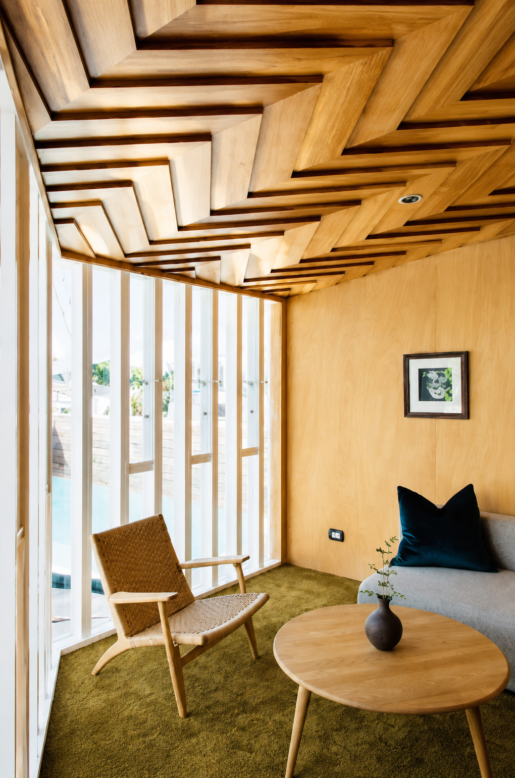 A modern interior room with a wooden zigzag ceiling, large glass windows, a wicker armchair, a small wooden coffee table with a dark vase, a beige sofa with a navy blue pillow, a green carpet, and a framed artwork on the wooden wall.