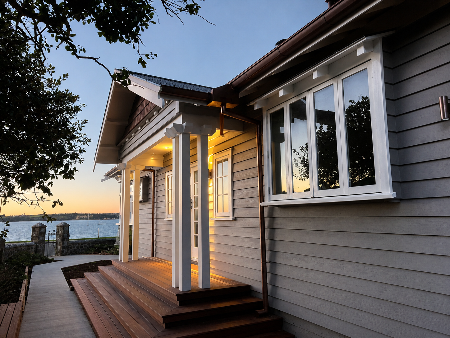 Exterior of a house near a body of water with a wooden porch and large windows during sunset.