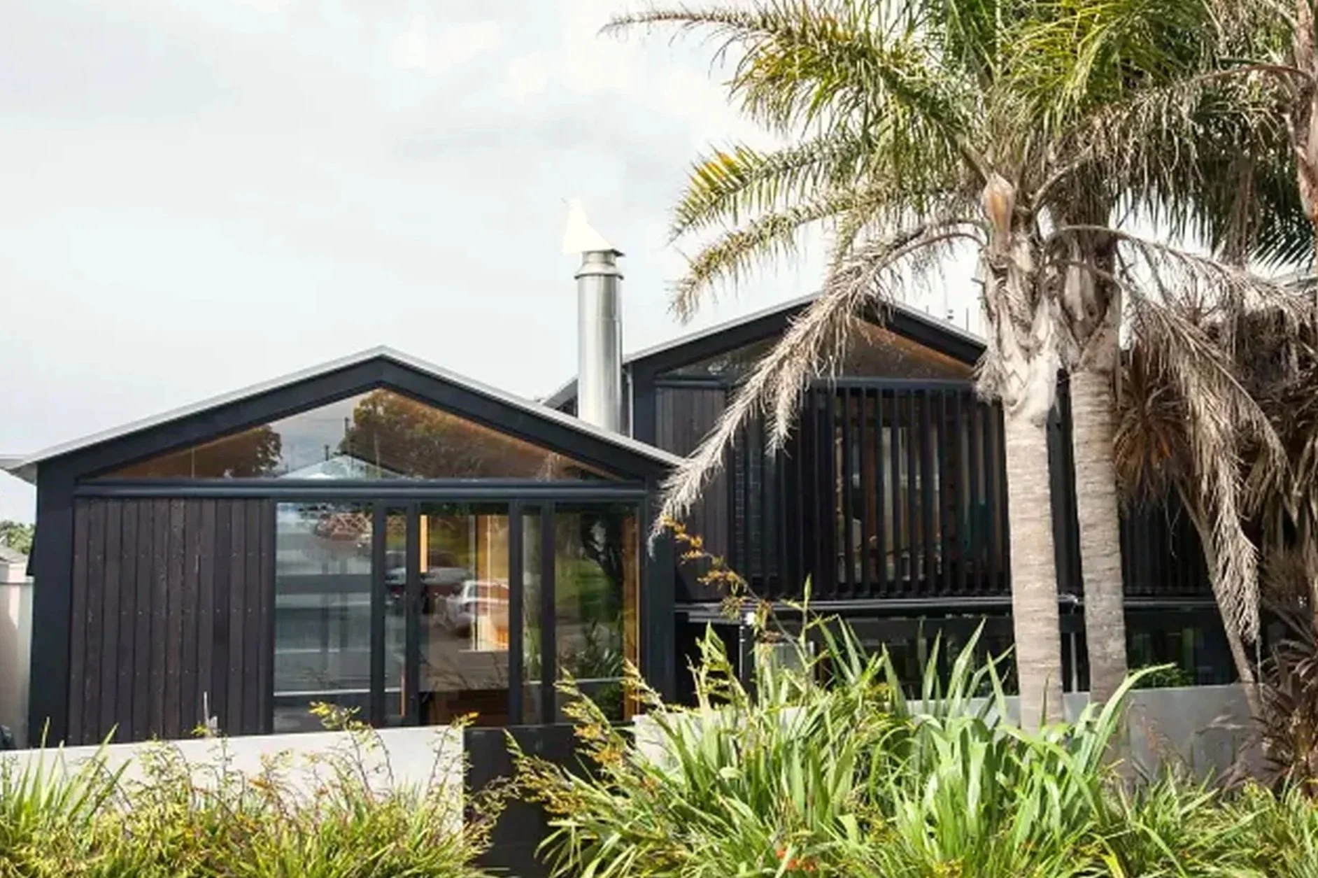 Modern black house with large glass windows and a sloped roof, surrounded by green plants and tall palms.