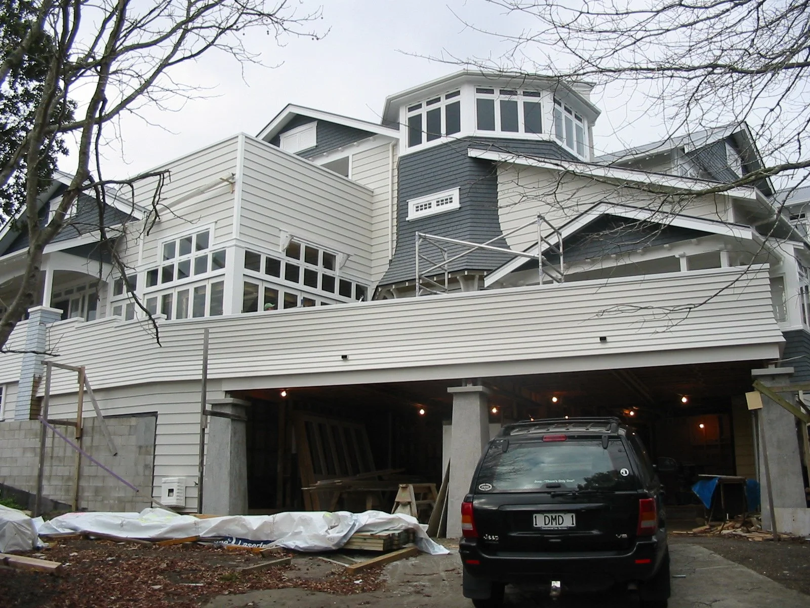 Under construction house with multiple levels and large windows, a black SUV parked in the driveway, construction materials and tools scattered on the ground below, and trees with bare branches in the foreground.