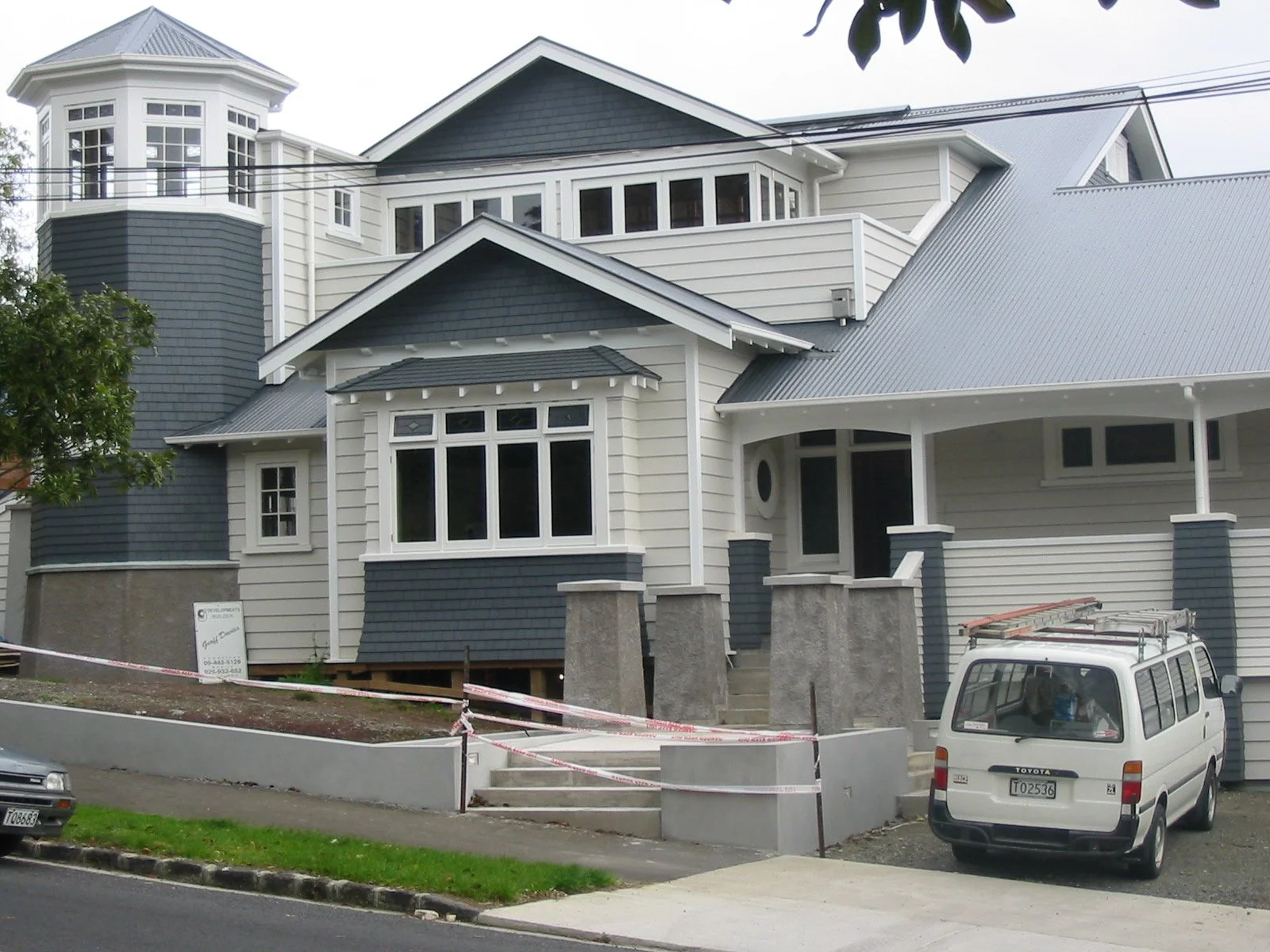 A large, multi-story house with a combination of white and dark blue siding and a gray metal roof. The house has multiple windows, including a prominent bay window and a corner tower with large windows. There is a front porch with steps and concrete 