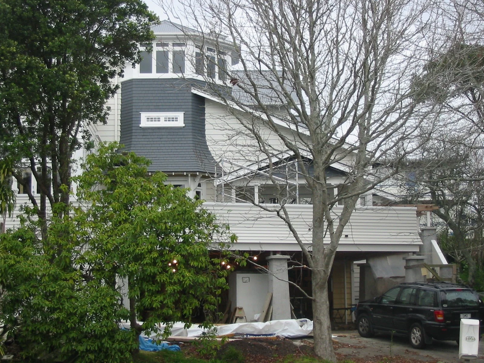A large multi-story house with a blue shingled roof, white siding, and a small window, partially obscured by leafless trees in the front yard, with a black car parked in the driveway and construction materials nearby.