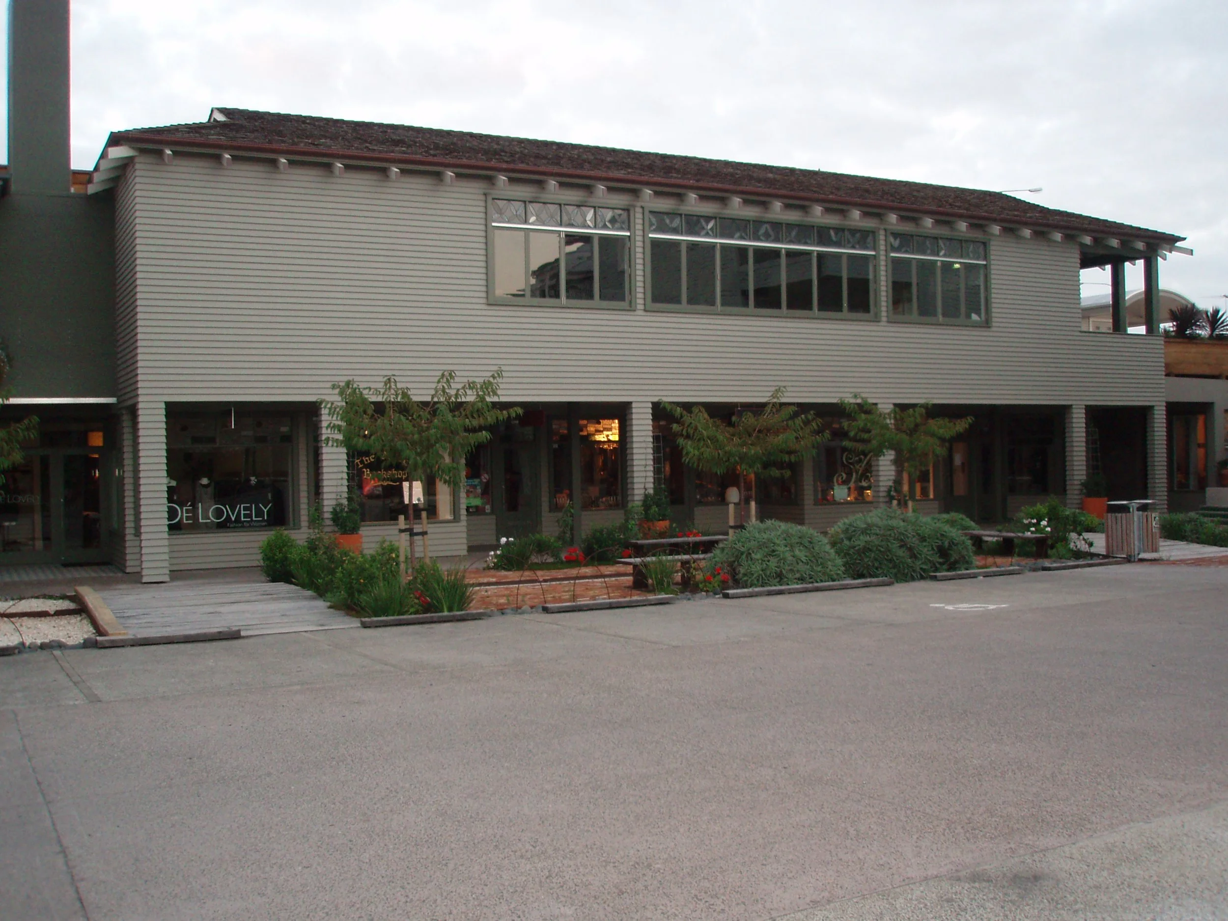 A two-story building with beige siding, large windows, and a storefront on the ground level. The storefront has a sign that reads 'DÉ LOVELY' and is surrounded by small trees and plants. There is a parking lot in the foreground with no vehicles.