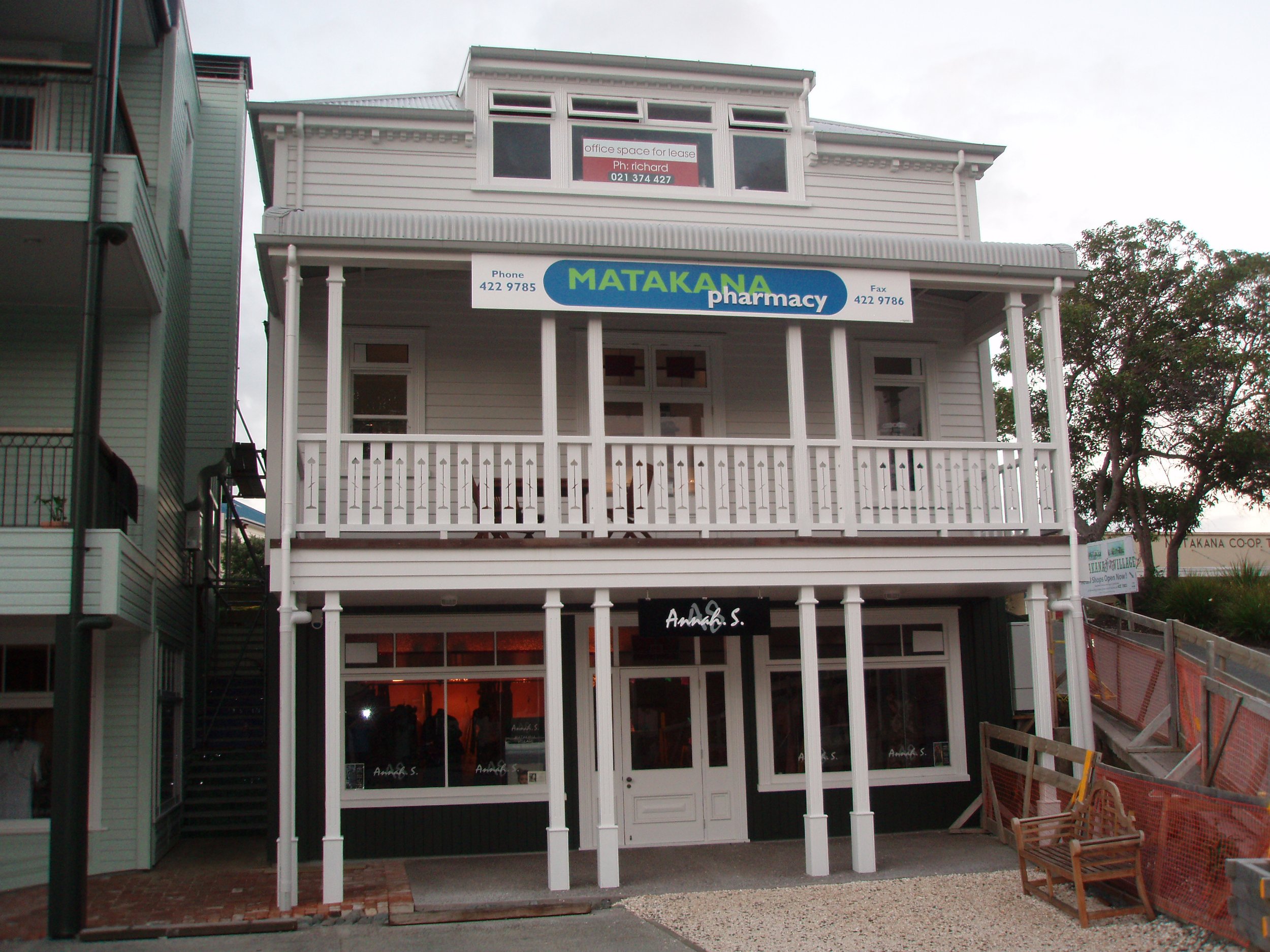 A multi-story building with a sign for Matakana Pharmacy on the front. The upper part has a sign indicating office space for lease with a contact number. The building has large glass windows and a white wooden balcony with a railing.